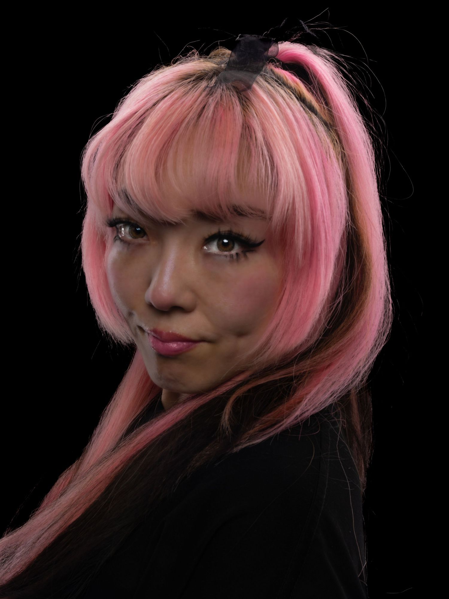 Young woman with pink and black hair, wearing a black top, posing against a dark background.
