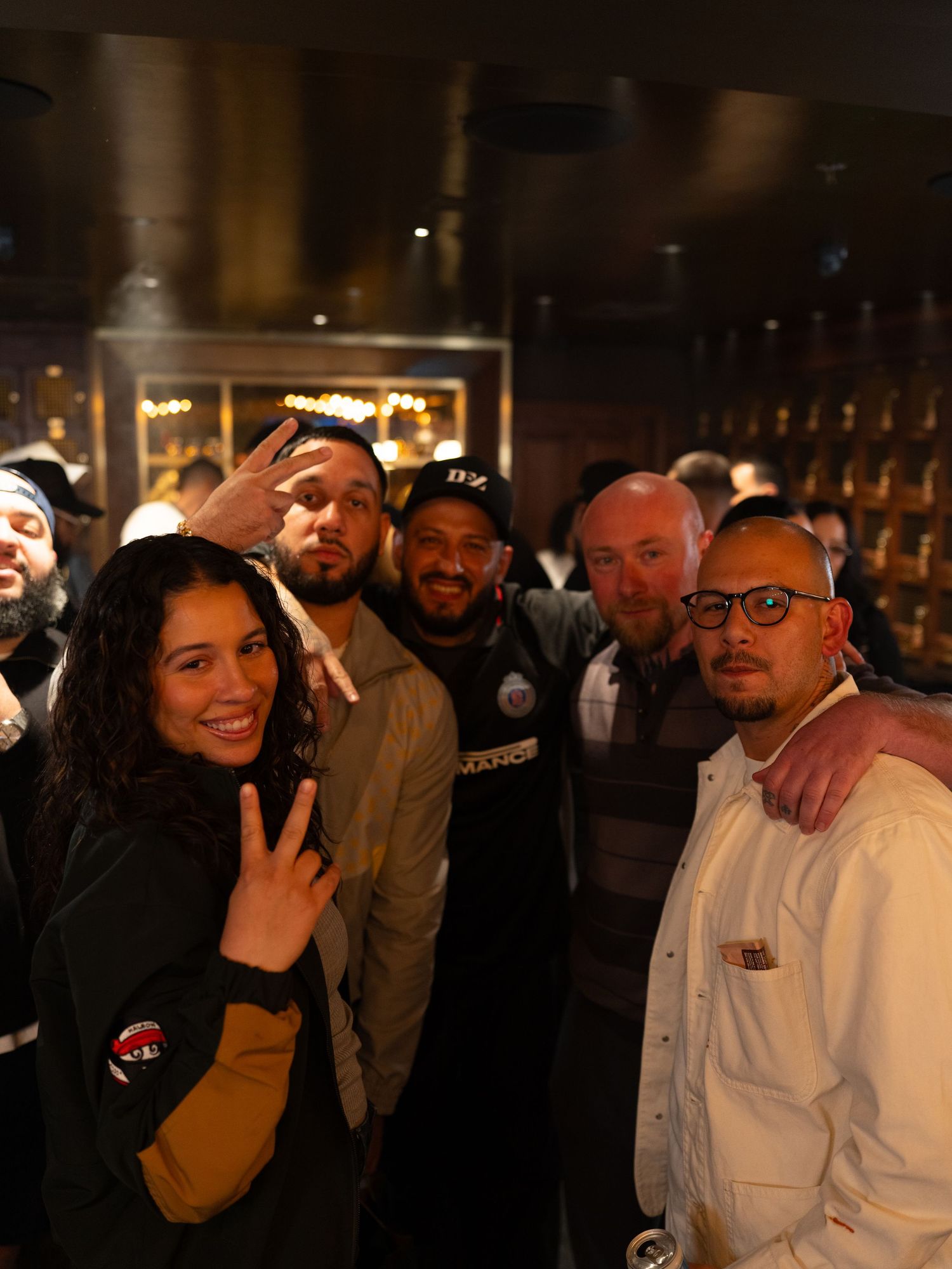 Group of friends posing together smiling at a dimly lit bar or lounge event, some flashing peace signs.
