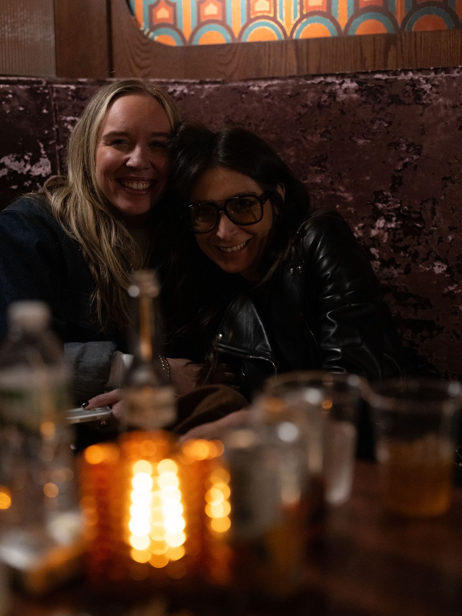 Two smiling women sitting together in a dimly lit bar booth with candles and drinks on the table.