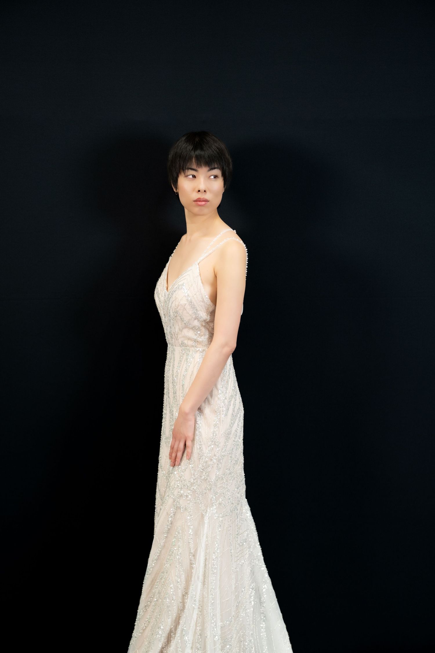 Model in flowing white lace dress poses against dark background in studio setting.