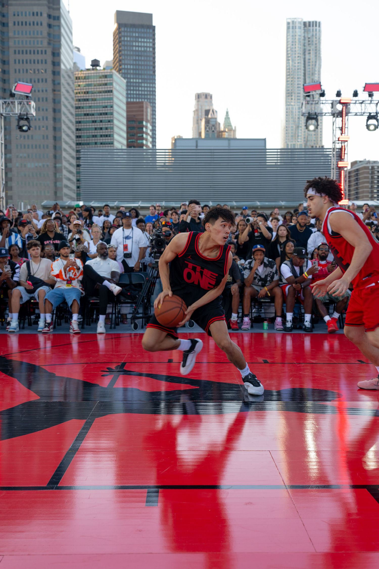 Basketball players competing on a red outdoor court with city skyscrapers in the background.