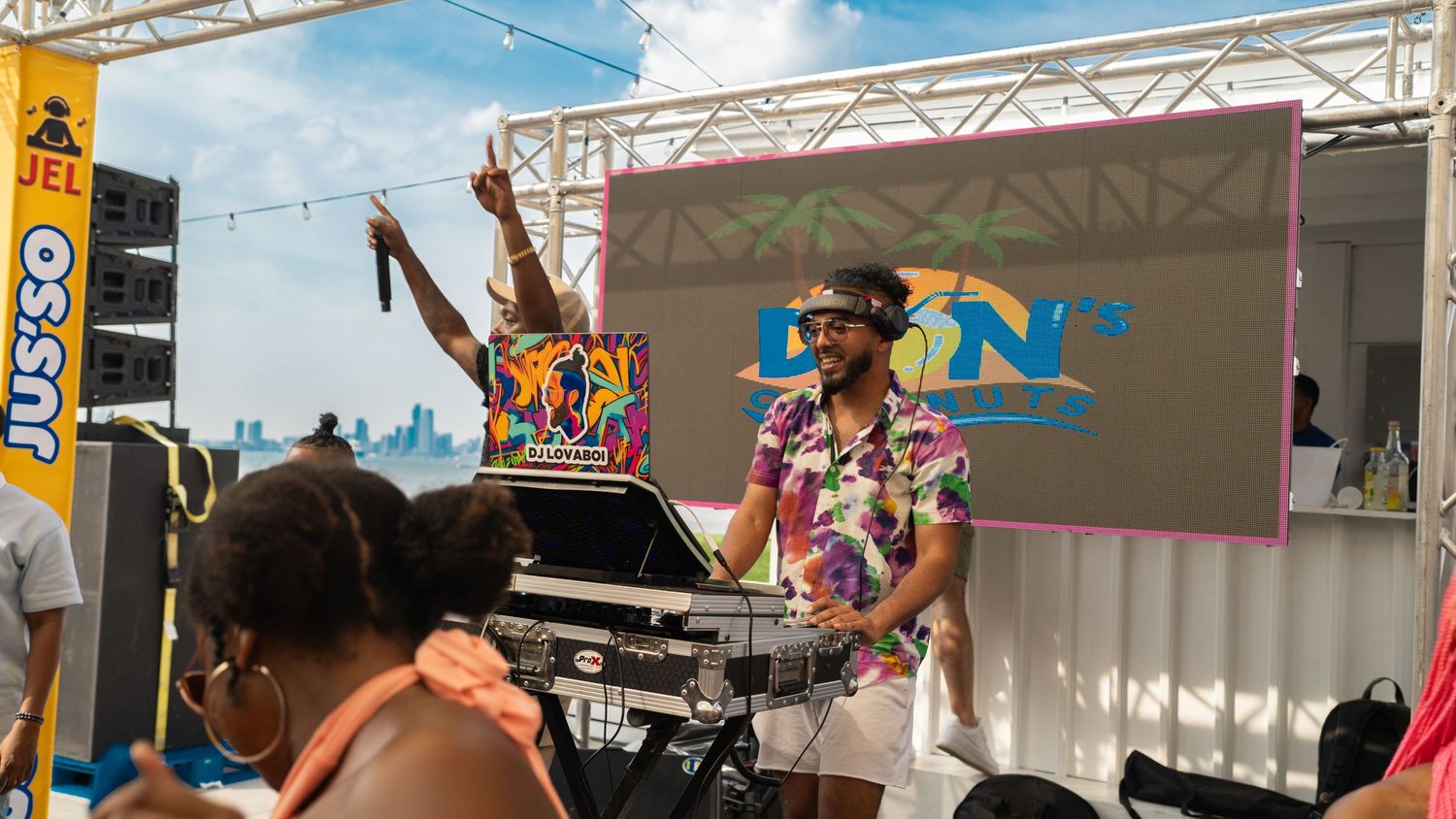DJ performing on stage with turntables at a daytime beach festival.