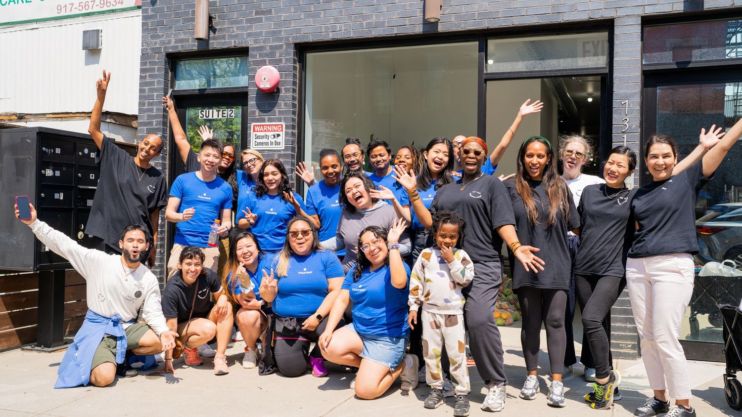 Large group of people posing together outside a brick building with raised arms in celebration.