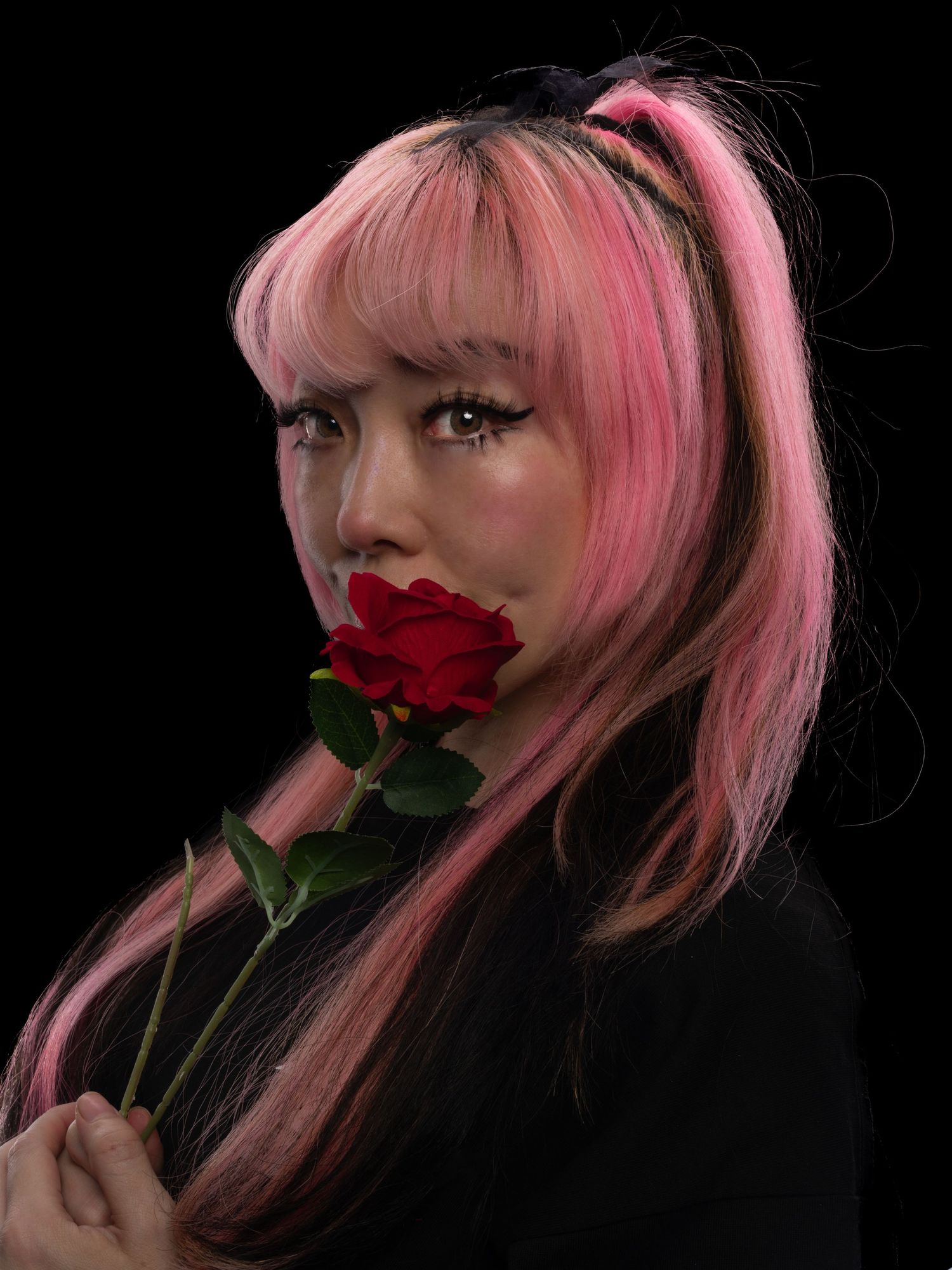 Young woman with pink and black hair holding a red rose near her lips against a dark background.
