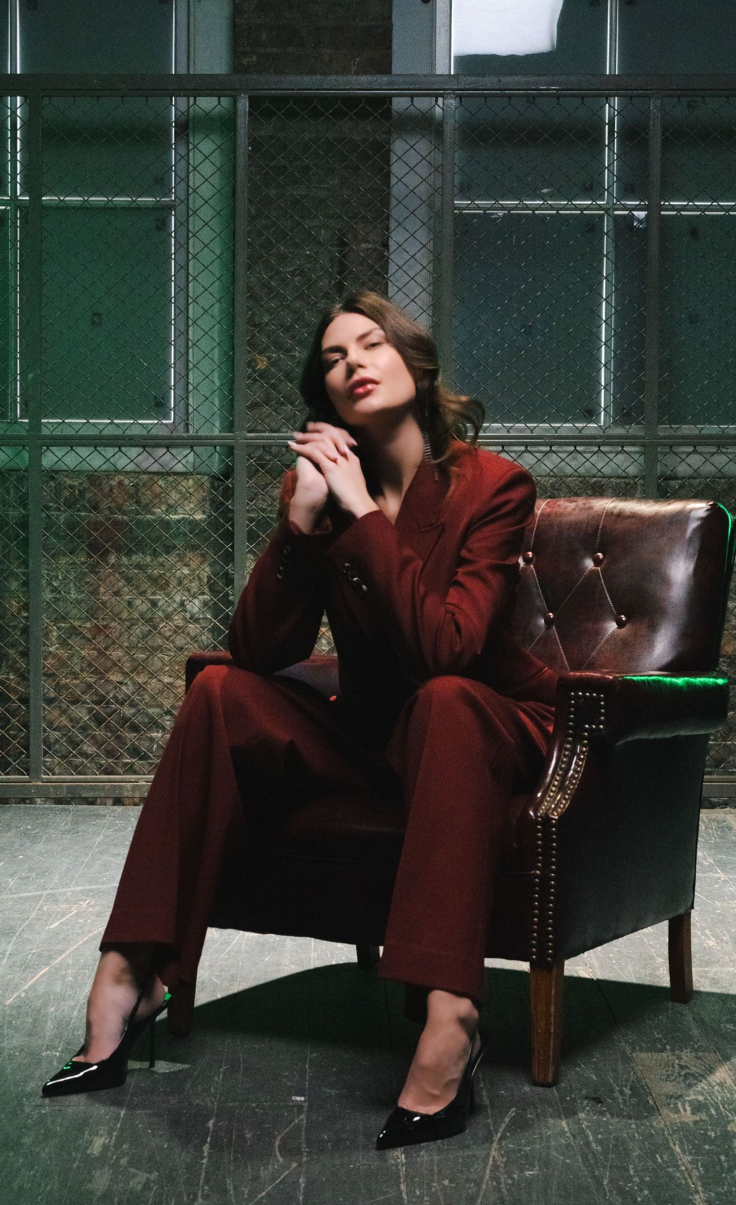 Model in burgundy suit lounging dramatically on leather couch in industrial setting.
