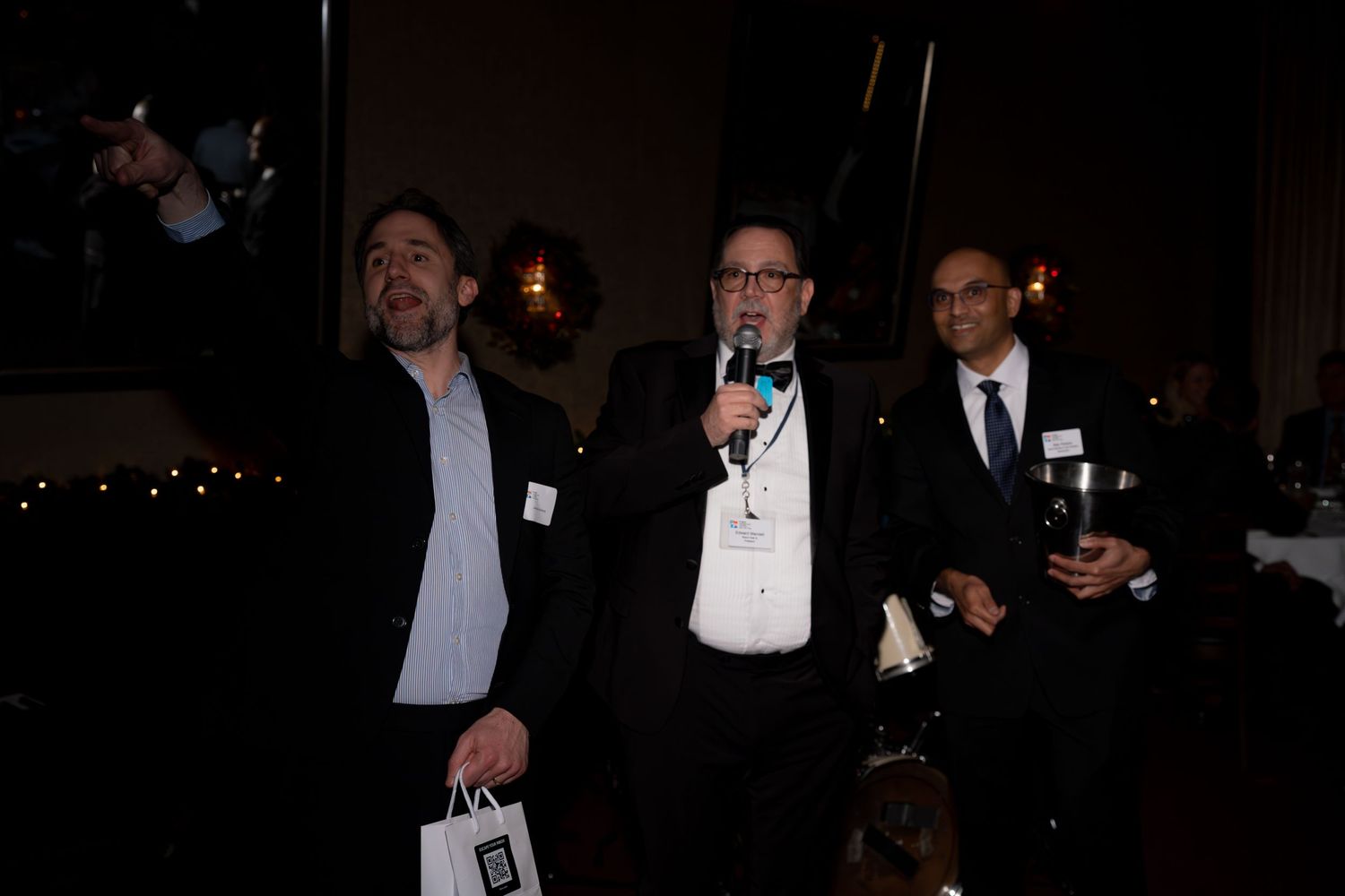 A group of formally dressed people in black tuxedos and evening wear gathered at a dimly lit social event.