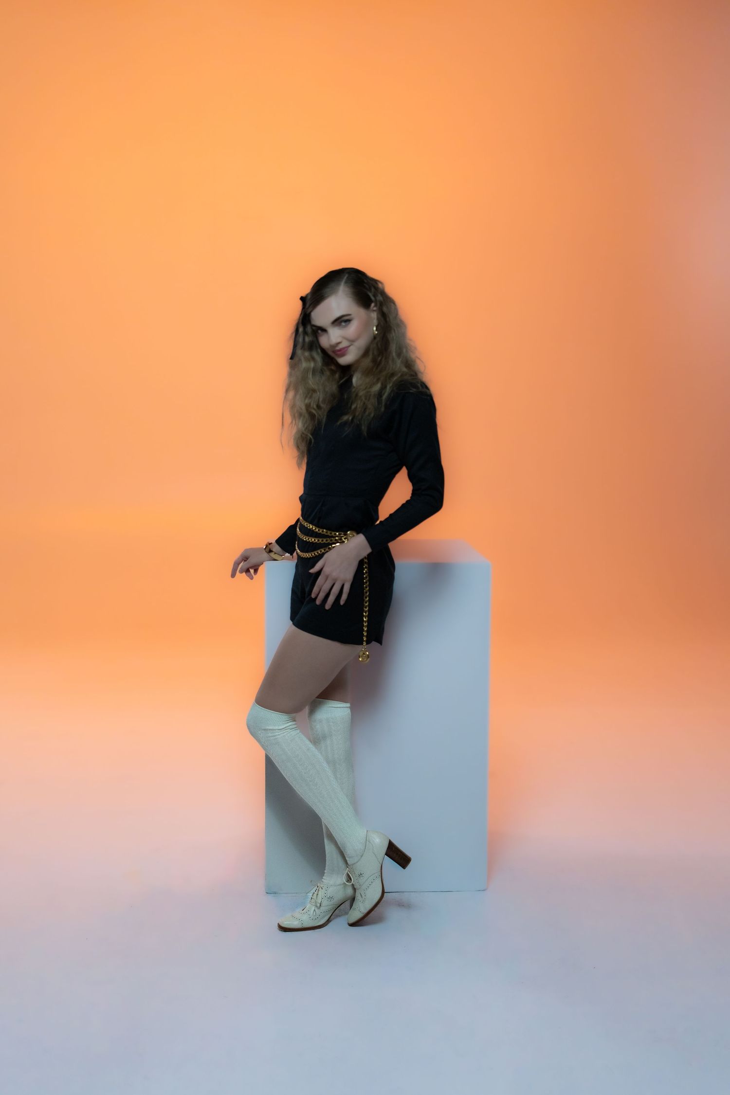 Model wearing black top and white boots poses on cube against orange gradient background.