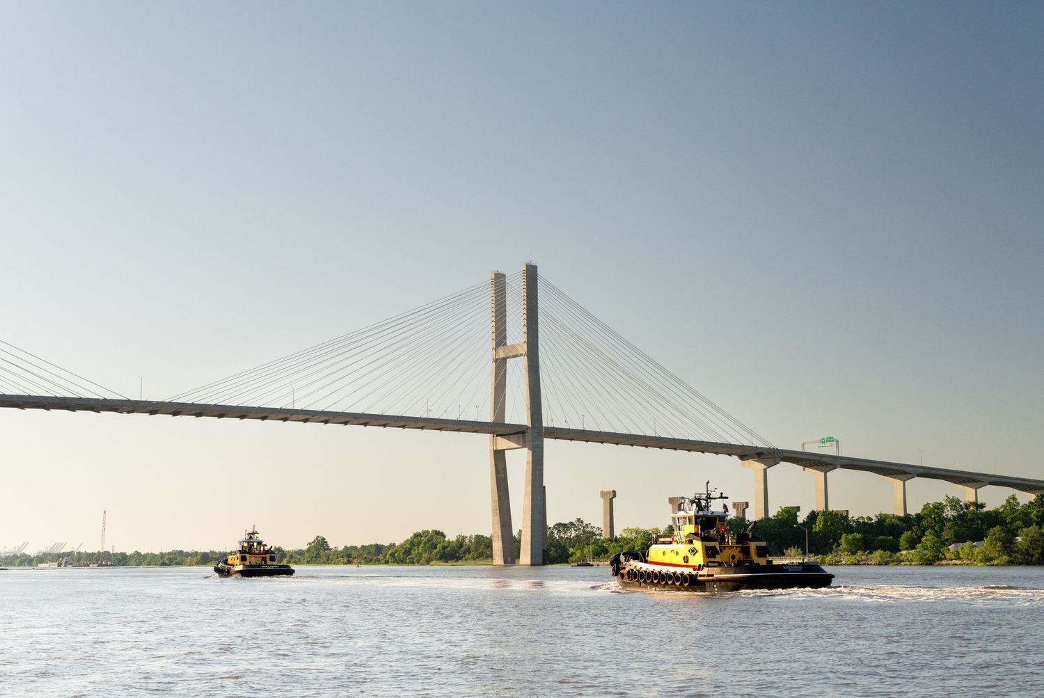 Cable suspension bridge spanning across a wide river during sunset.