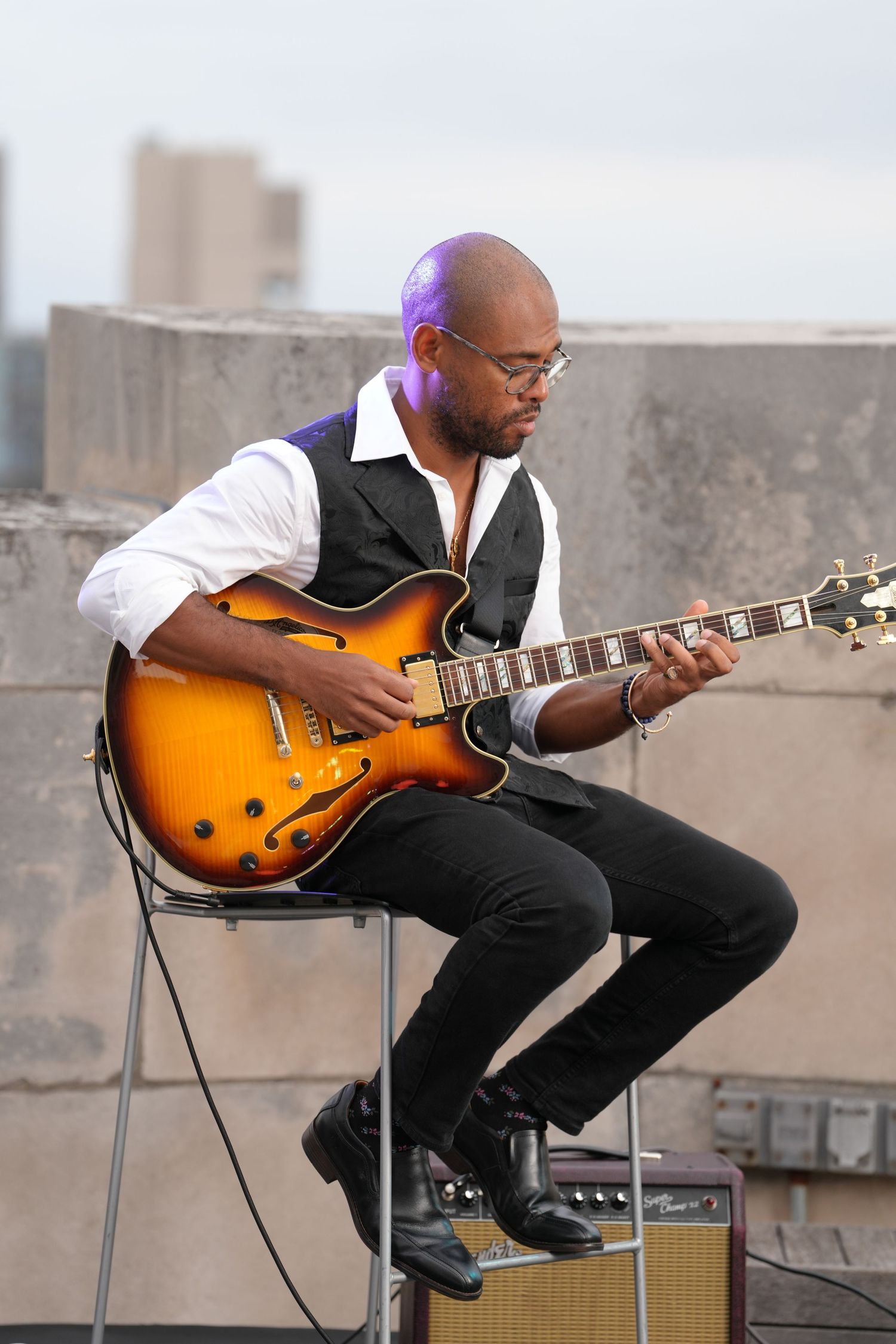 Musician plays a sunburst electric guitar while seated on a rooftop with urban buildings in the background.