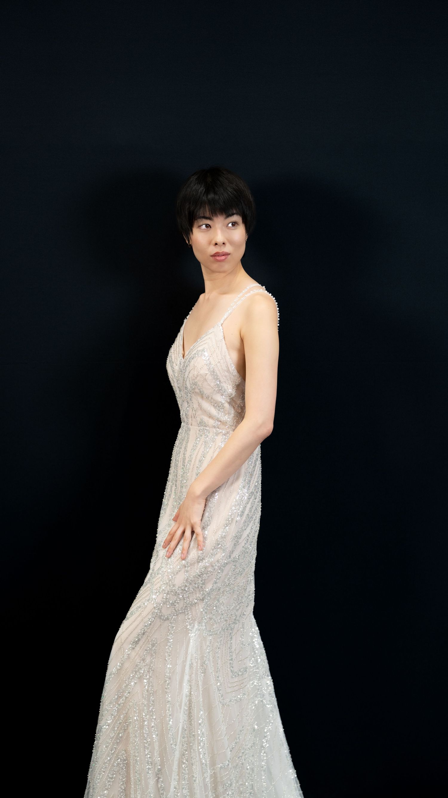 Person wearing flowing white sequined gown poses elegantly against black backdrop.