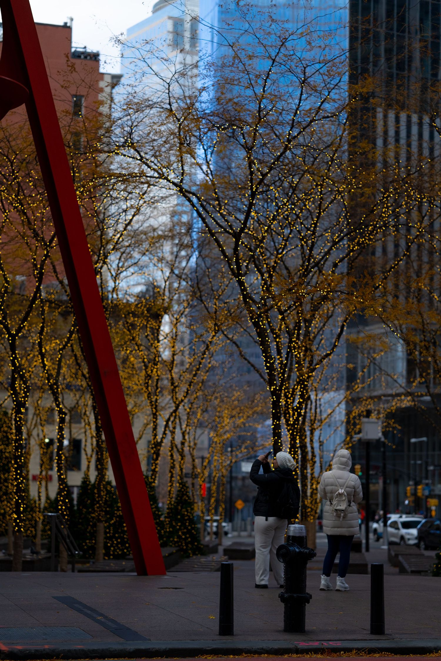 Urban street scene with tall buildings, autumn trees, and a large red structural beam in the foreground.