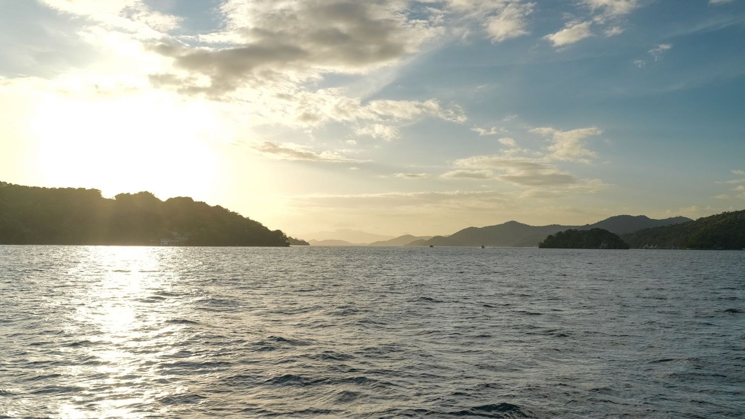 Panoramic sunset view over calm ocean waters with mountains silhouetted against a cloudy golden sky.