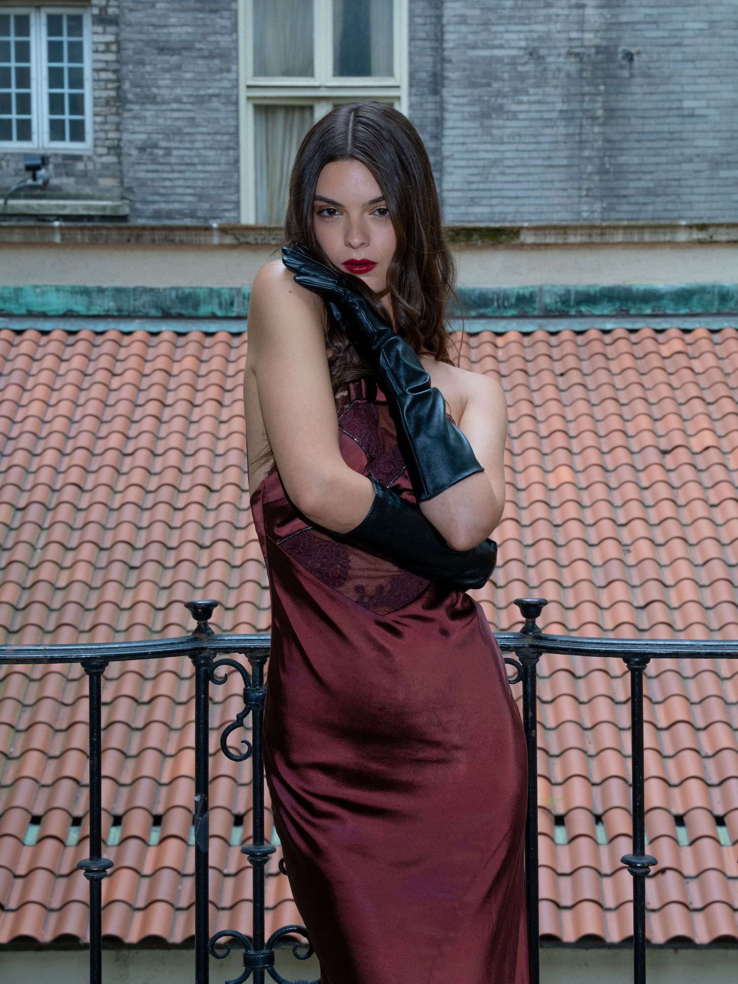 Woman in burgundy satin dress and black leather gloves poses on balcony with terracotta roof backdrop.