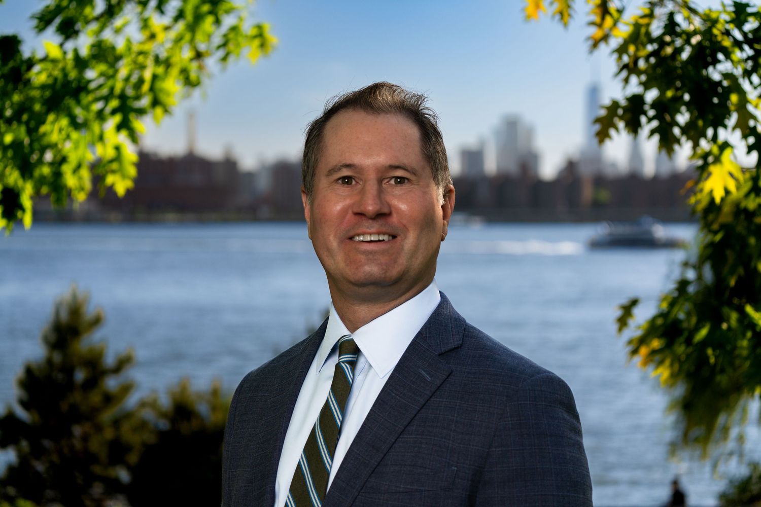 Outdoor business portrait with water and city skyline visible in background.