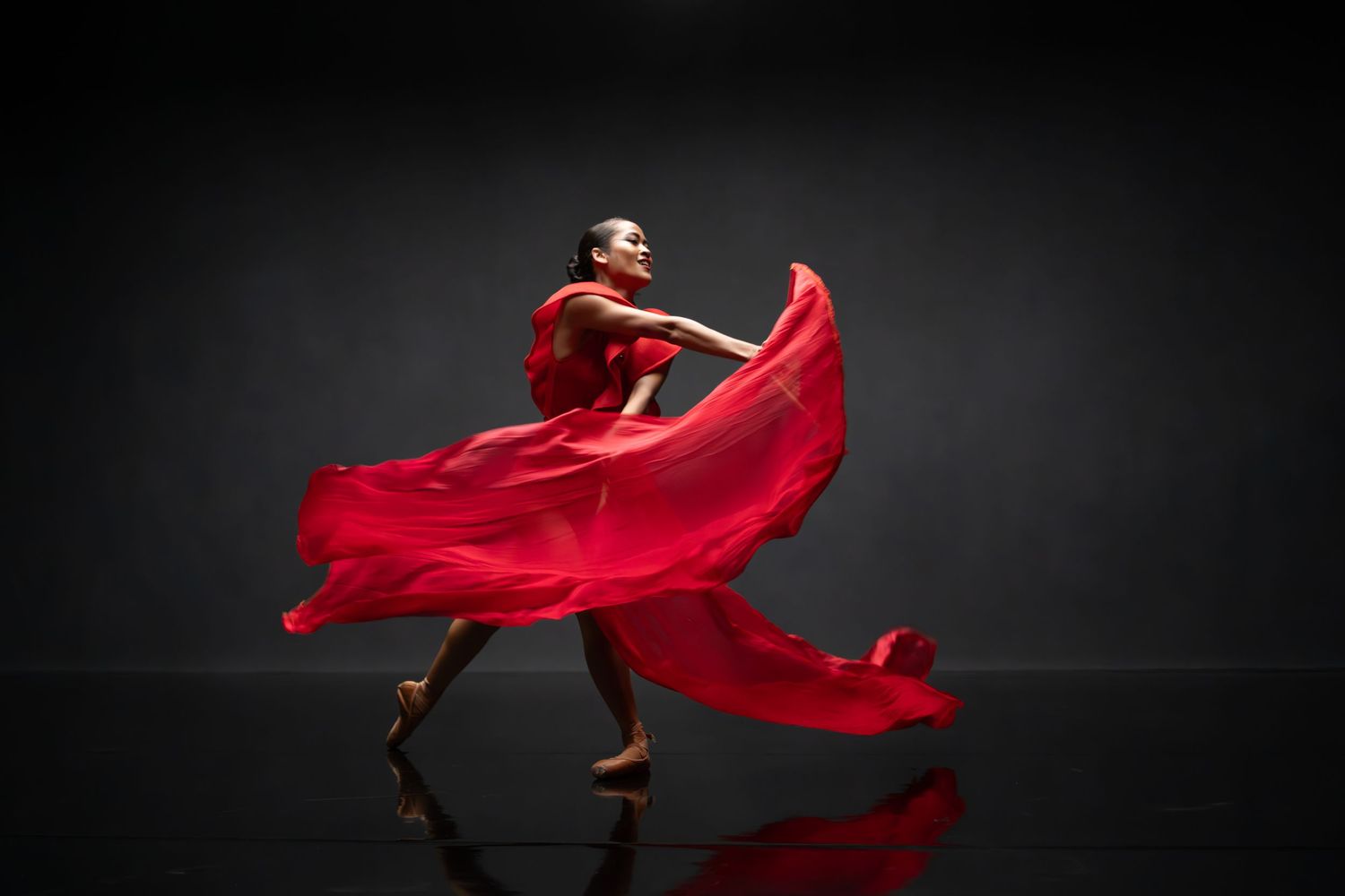 Performance artist in red flowing dress creates sweeping movement on dark stage.