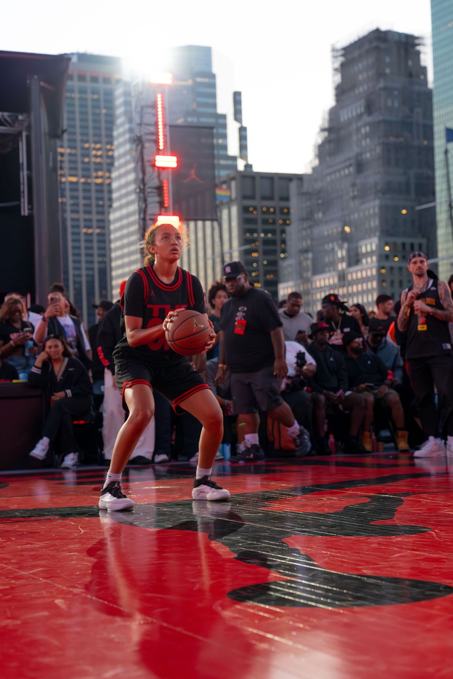 Athletes performing on red basketball court with city skyline backdrop at sunset.