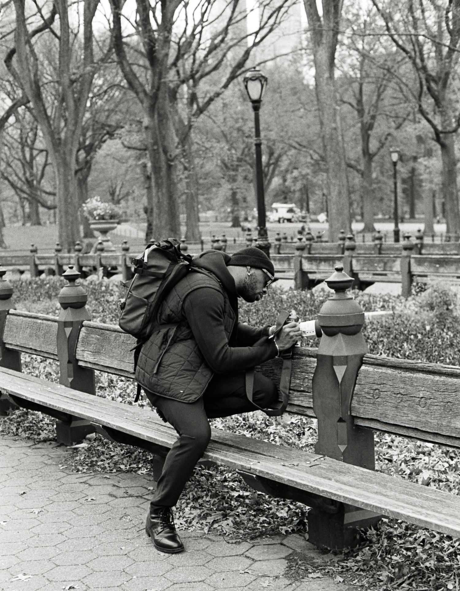 A person lies on a park bench in winter with fallen leaves scattered on the ground in this black and white photograph.