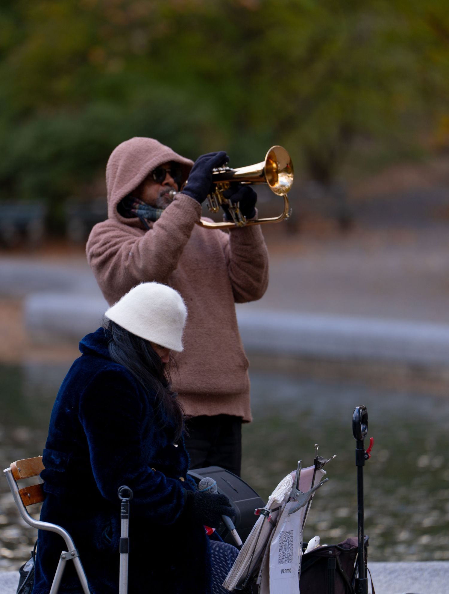 A person in a brown coat and white hat plays a trumpet outdoors in a series of sequential shots.