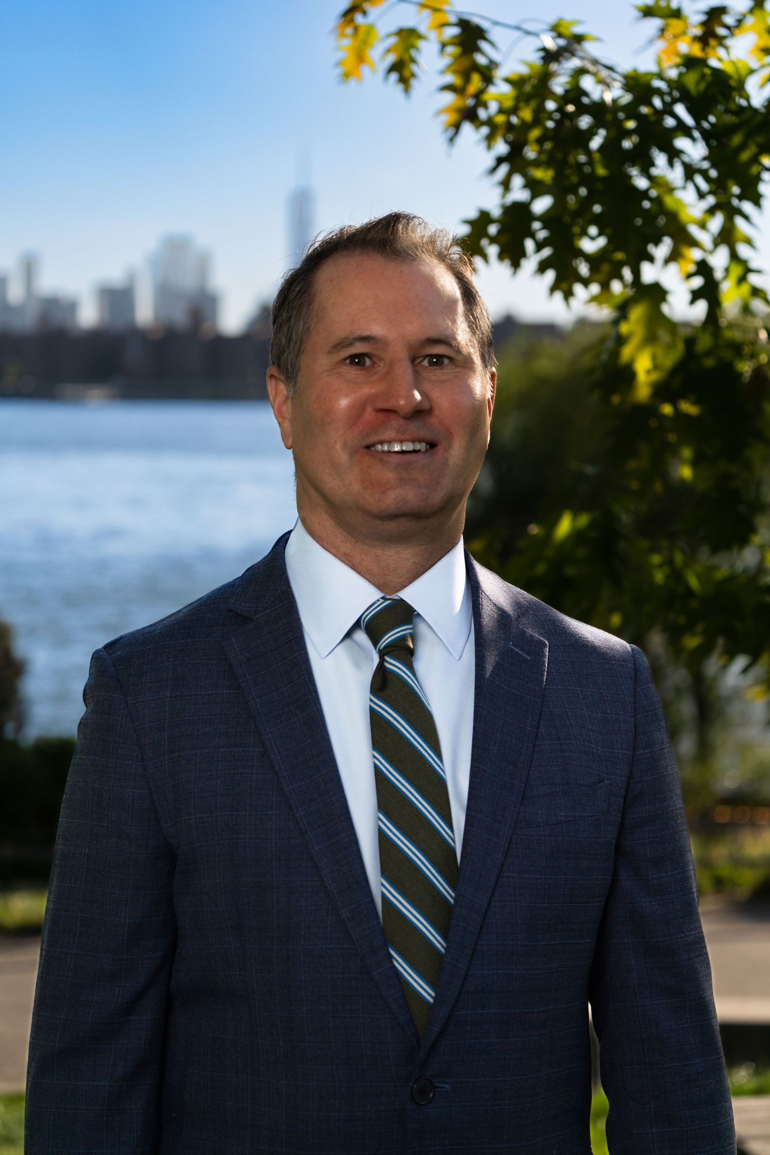 Professional portrait against waterfront with city skyline and trees in background.