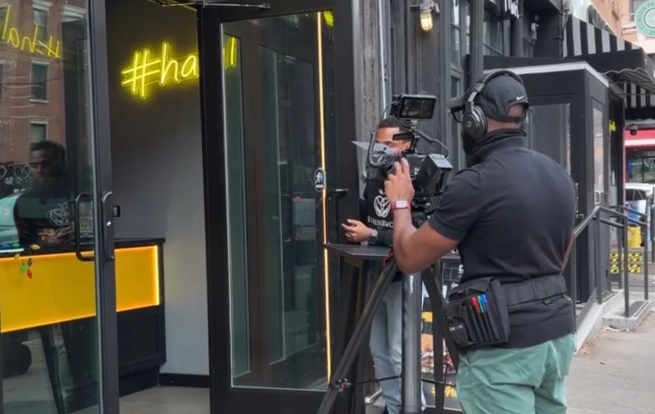 Camera crew filming outside a trendy urban restaurant with a yellow neon hashtag sign on the storefront.