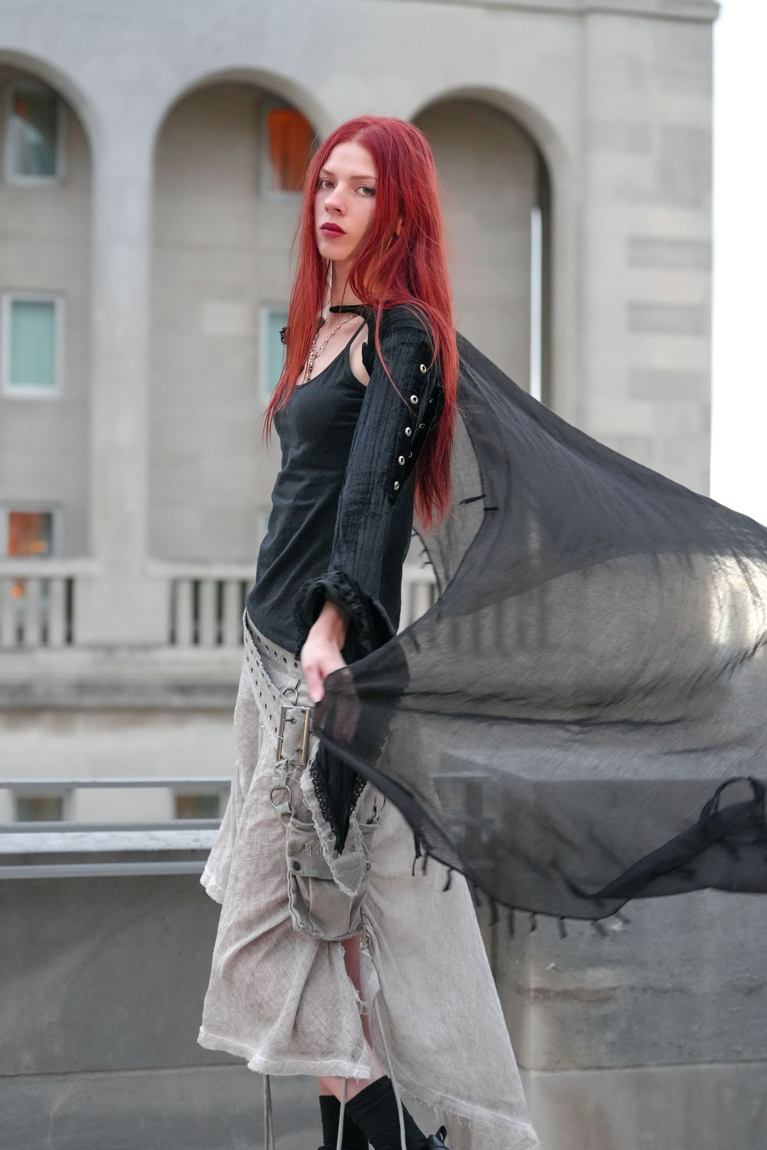 Fashion model with red hair twirls in black and gray outfit against architectural backdrop.