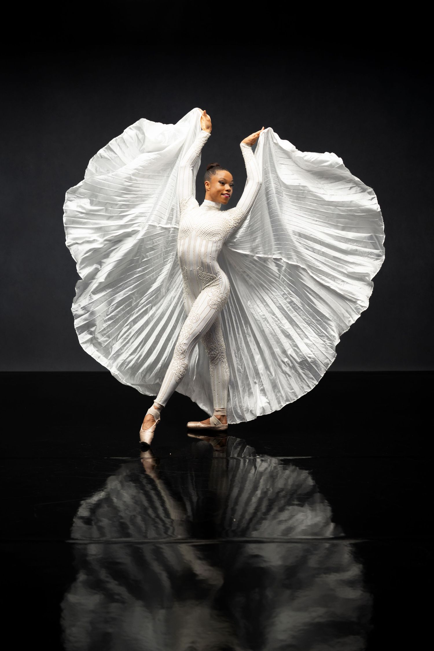 Dancer in flowing white dress performs graceful movements against dark background with floor reflection.