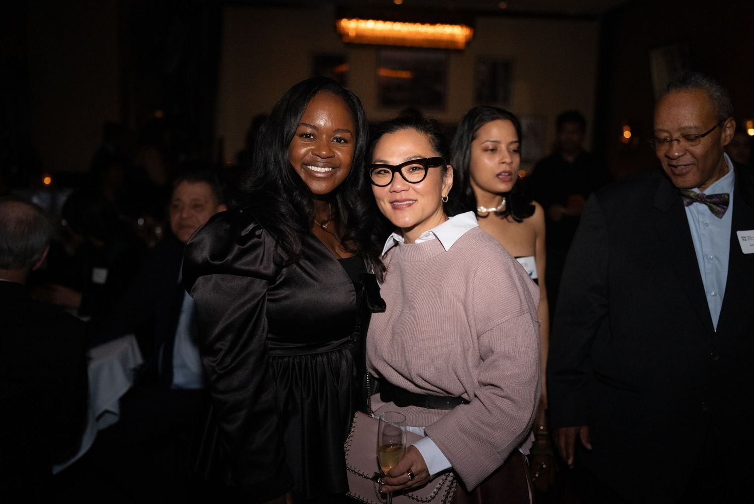 Guests in formal attire mingle and smile at an elegant evening event in a dimly lit venue.