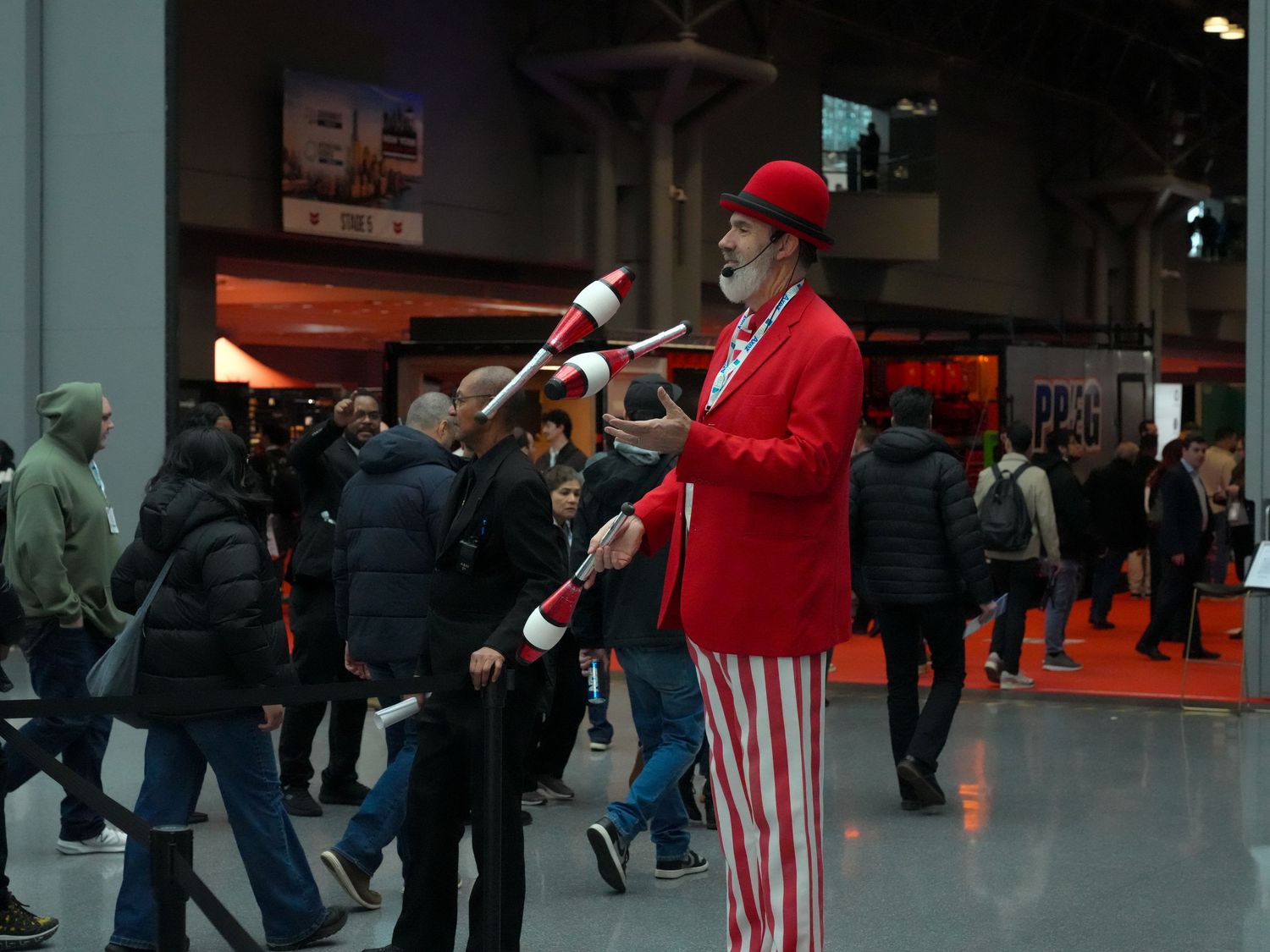 A clown in red suit and striped pants juggles pins at a busy indoor event or convention center.
