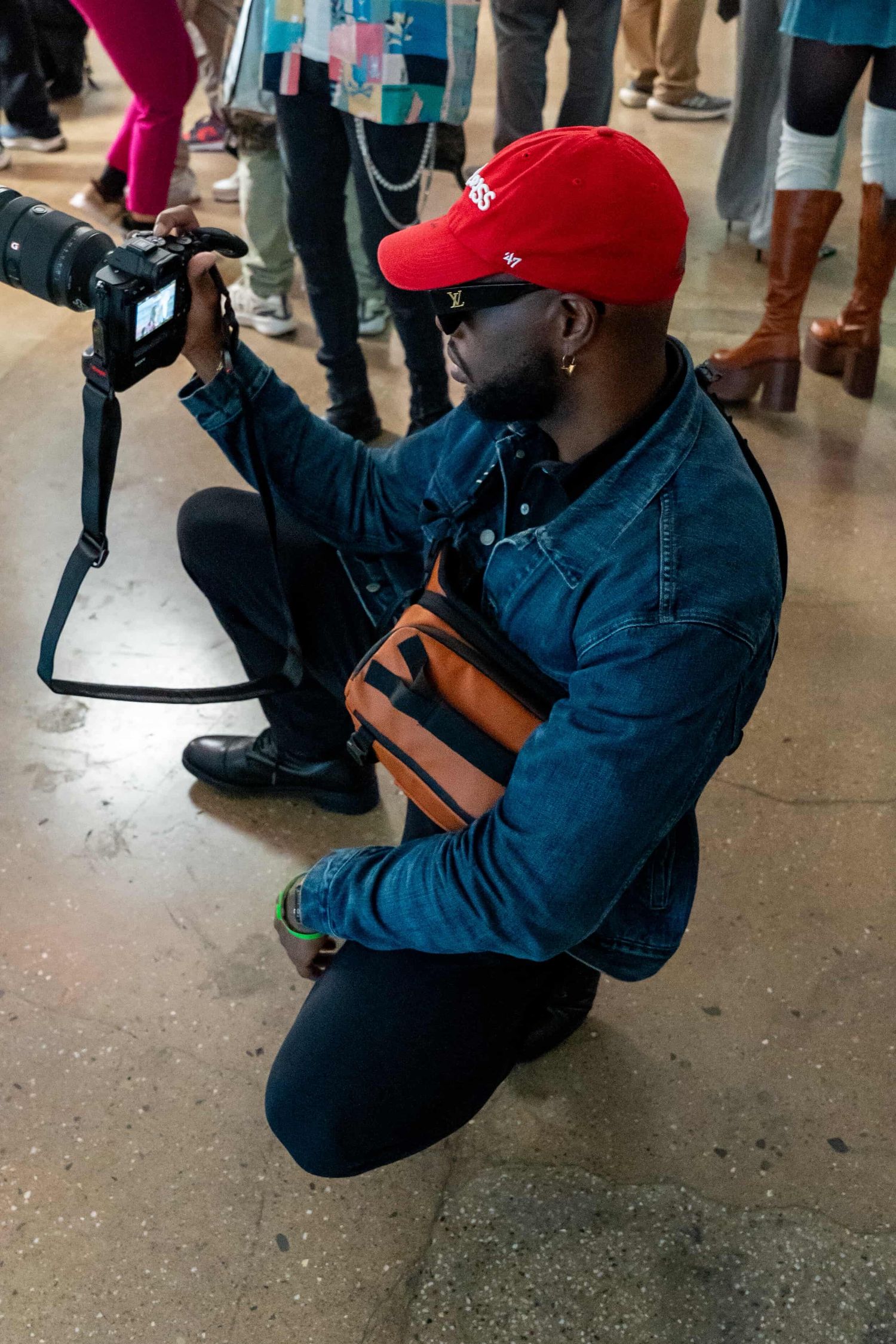 A photographer in a red baseball cap and navy jacket crouches low to capture a shot on their professional camera.