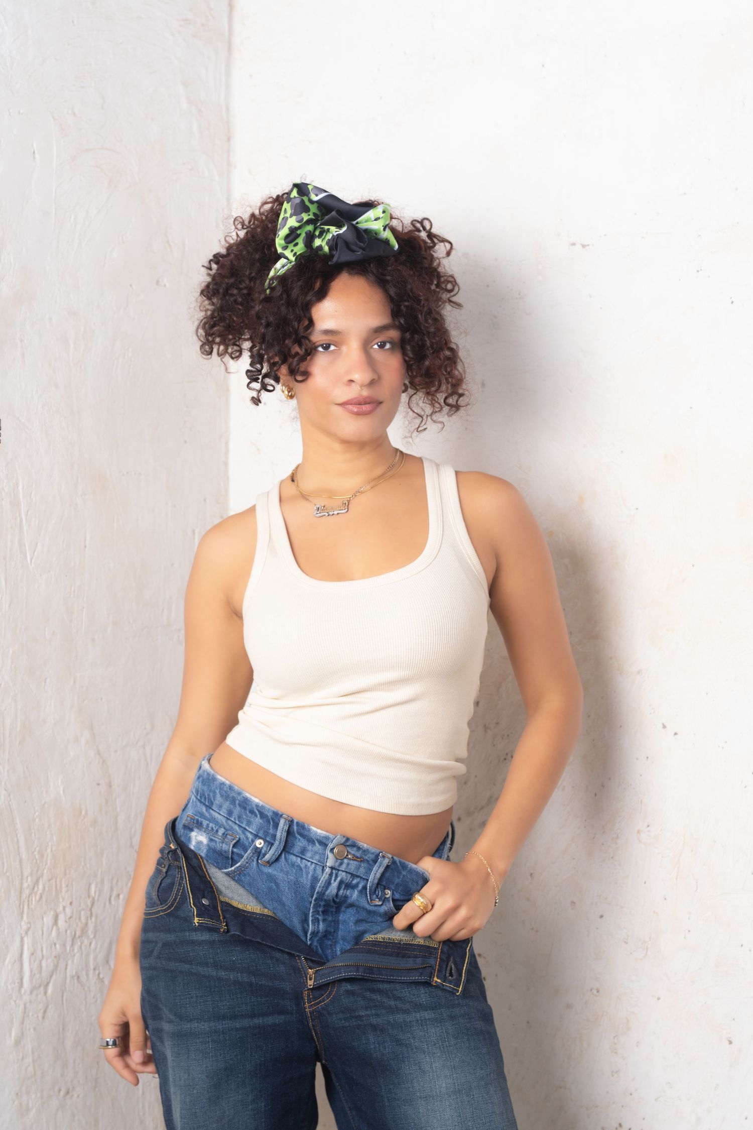Person in white tank top and blue jeans poses against white wall in a casual fashion photoshoot series.
