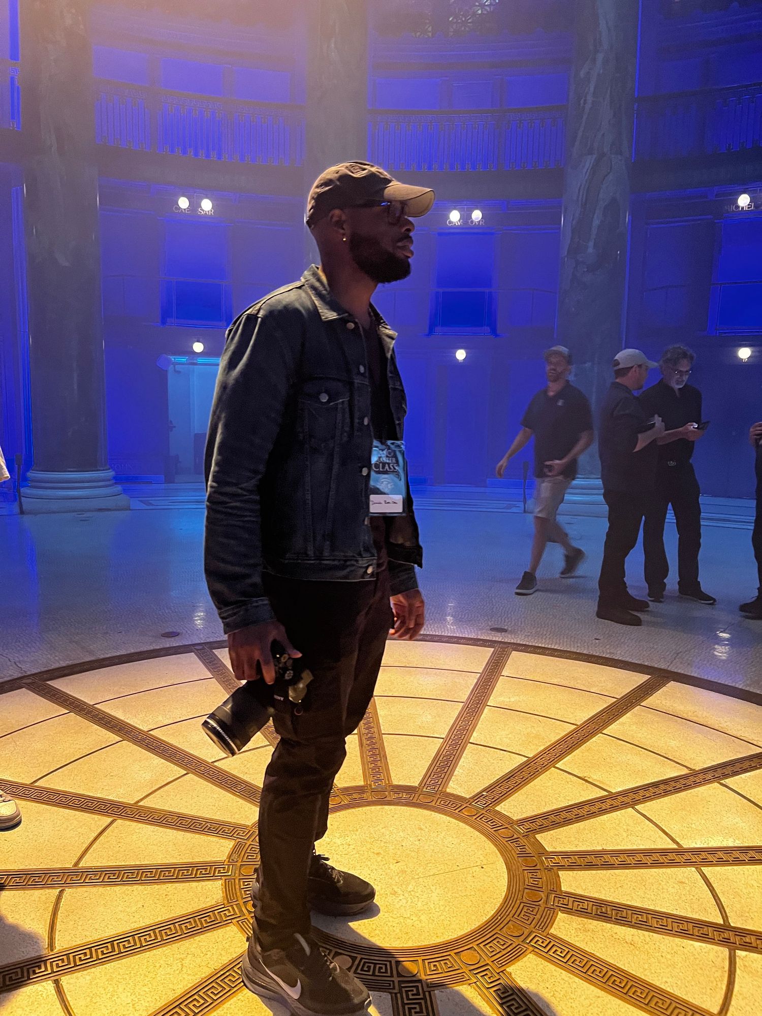 Man in denim jacket and cap stands on ornate gold floor in blue-lit columned hall holding a camera.