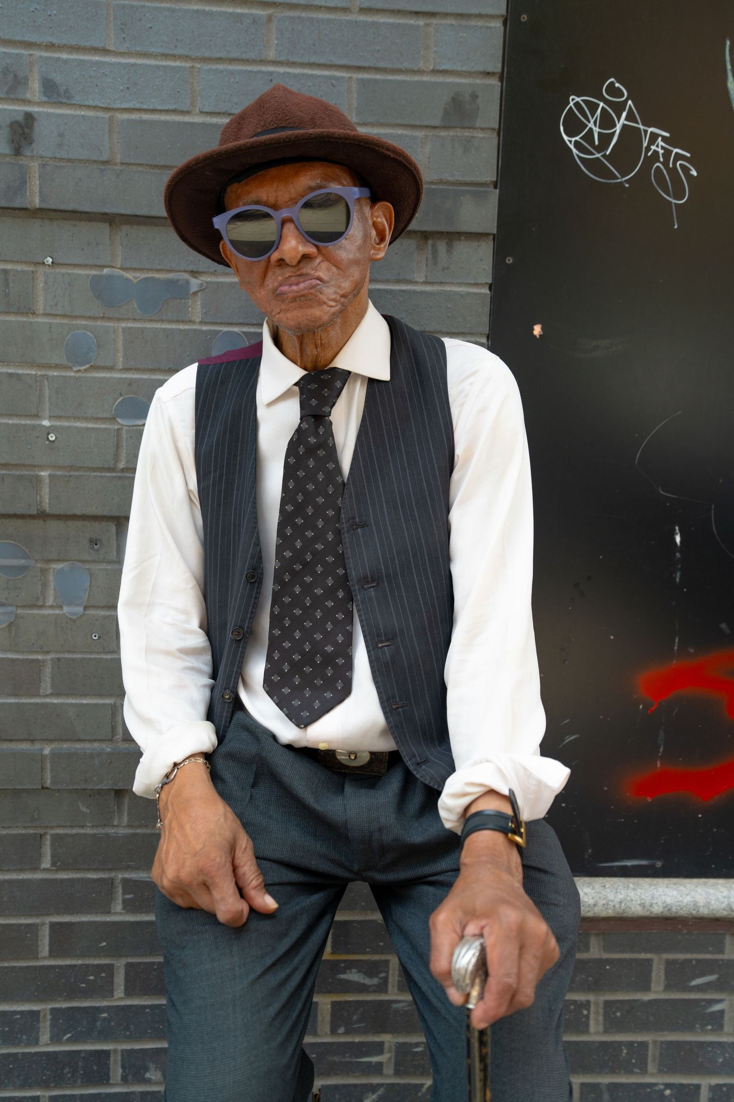 A person in formal attire with pinstripe vest and brown hat poses against an urban backdrop.
