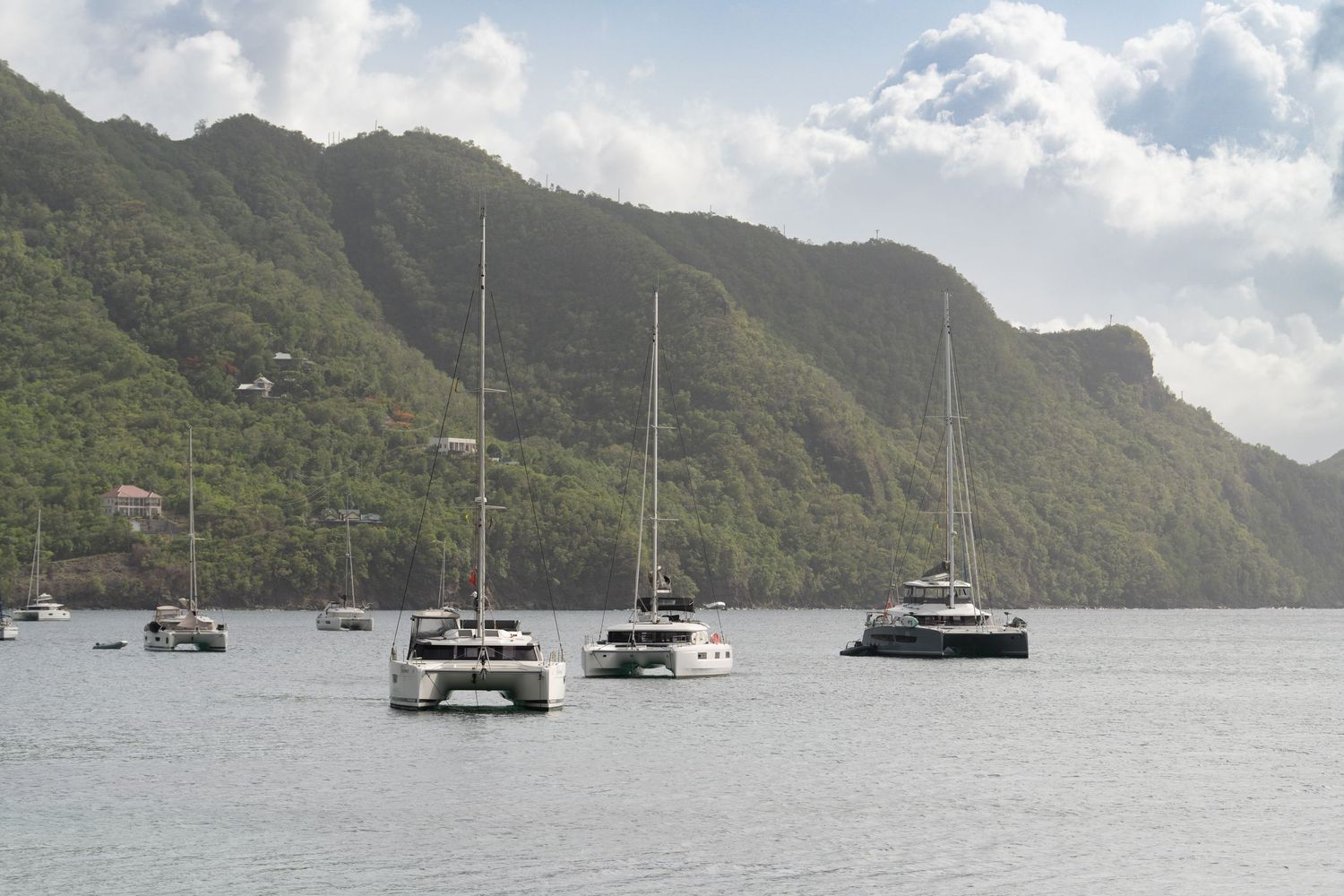 Luxury catamarans and yachts moored in Caribbean harbor surrounded by dramatic mountain landscape.