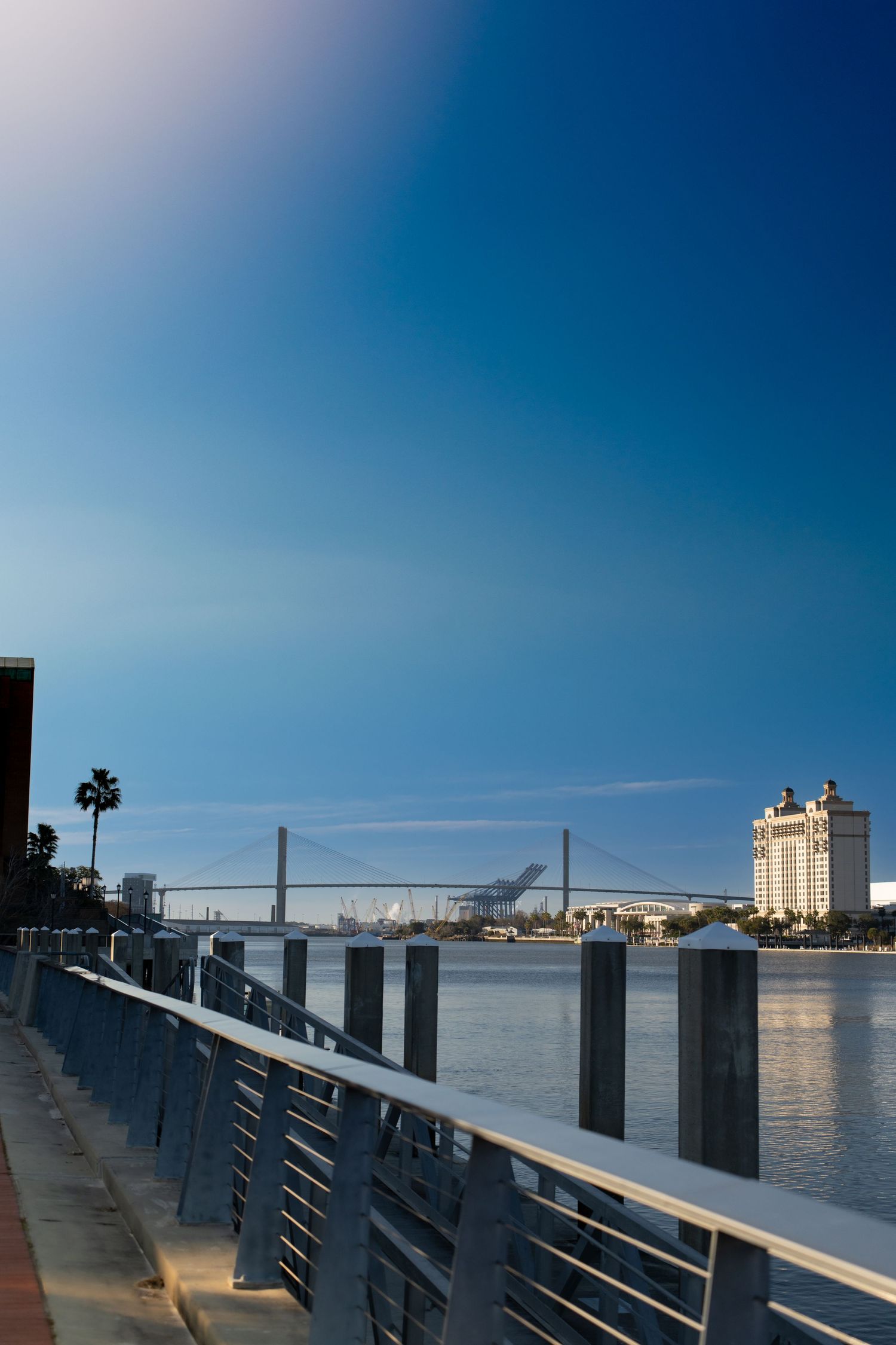 Waterfront boardwalk with metal railings overlooking a river with bridge and buildings in the distance.