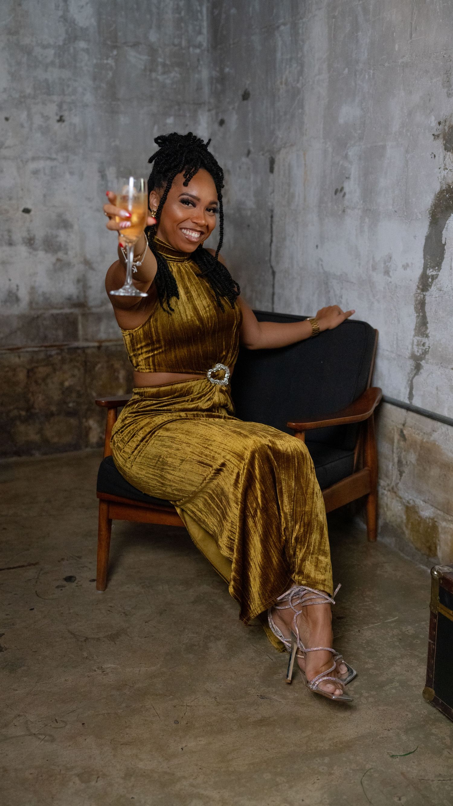 Someone in a gold velvet dress lounges on a vintage chair while holding a wine glass against a concrete wall.