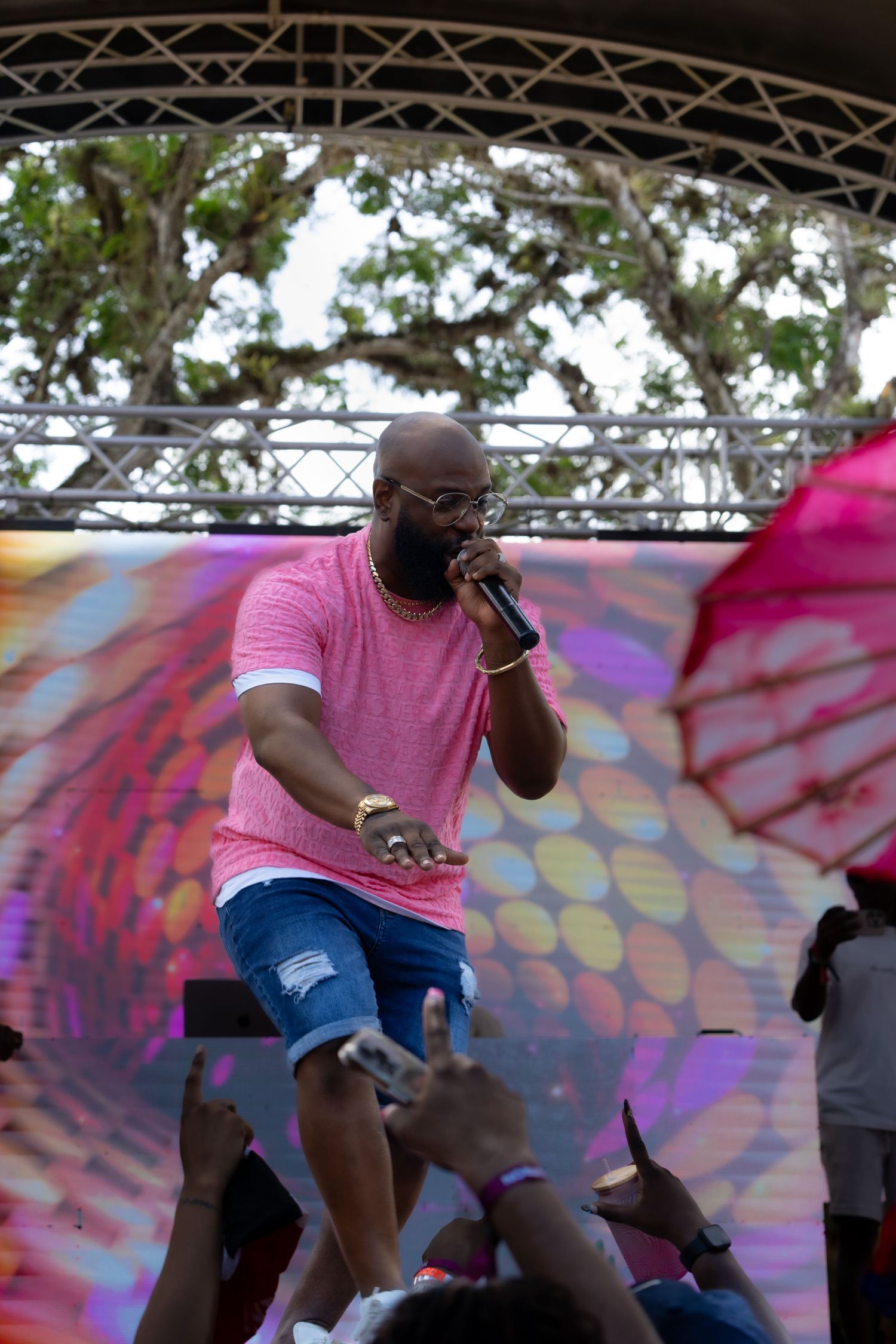 Performer in pink shirt and denim shorts singing on outdoor festival stage.