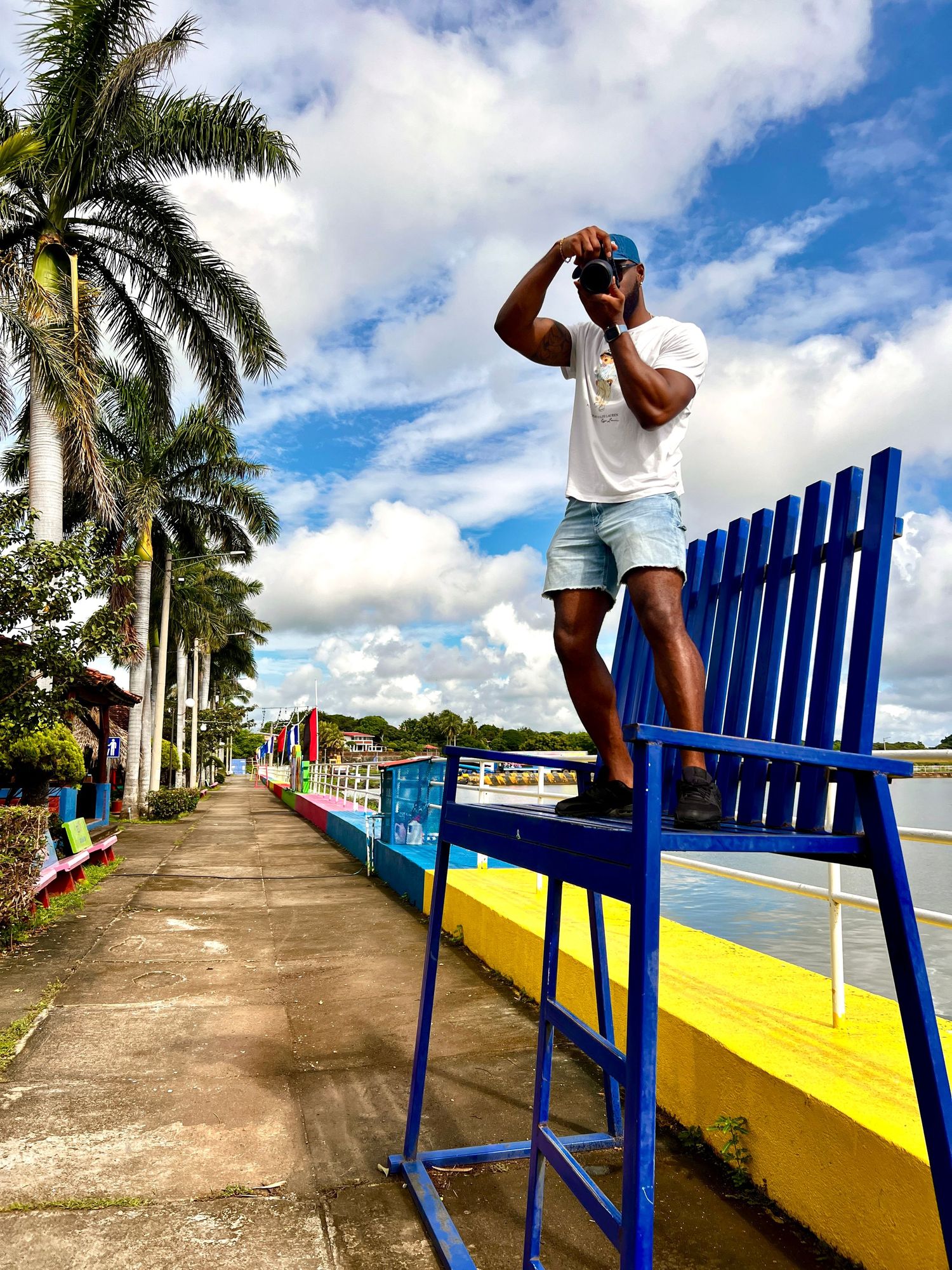 Bright blue bench along palm tree-lined waterfront walkway under cloudy sky.
