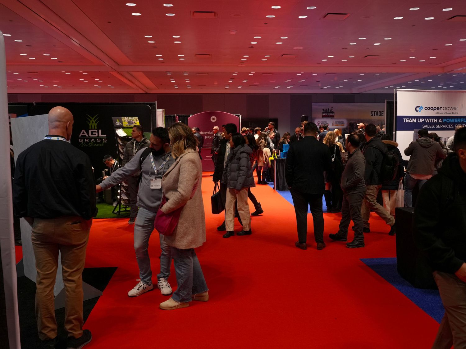 Busy trade show floor with attendees networking near vendor booths including AGL, on red carpet under pink lighting.