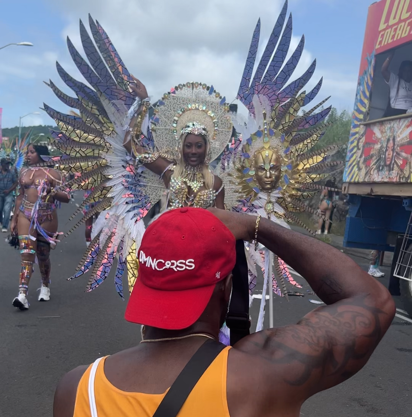 Person in red cap photographing elaborate feathered carnival costume with golden wings at a vibrant street parade.