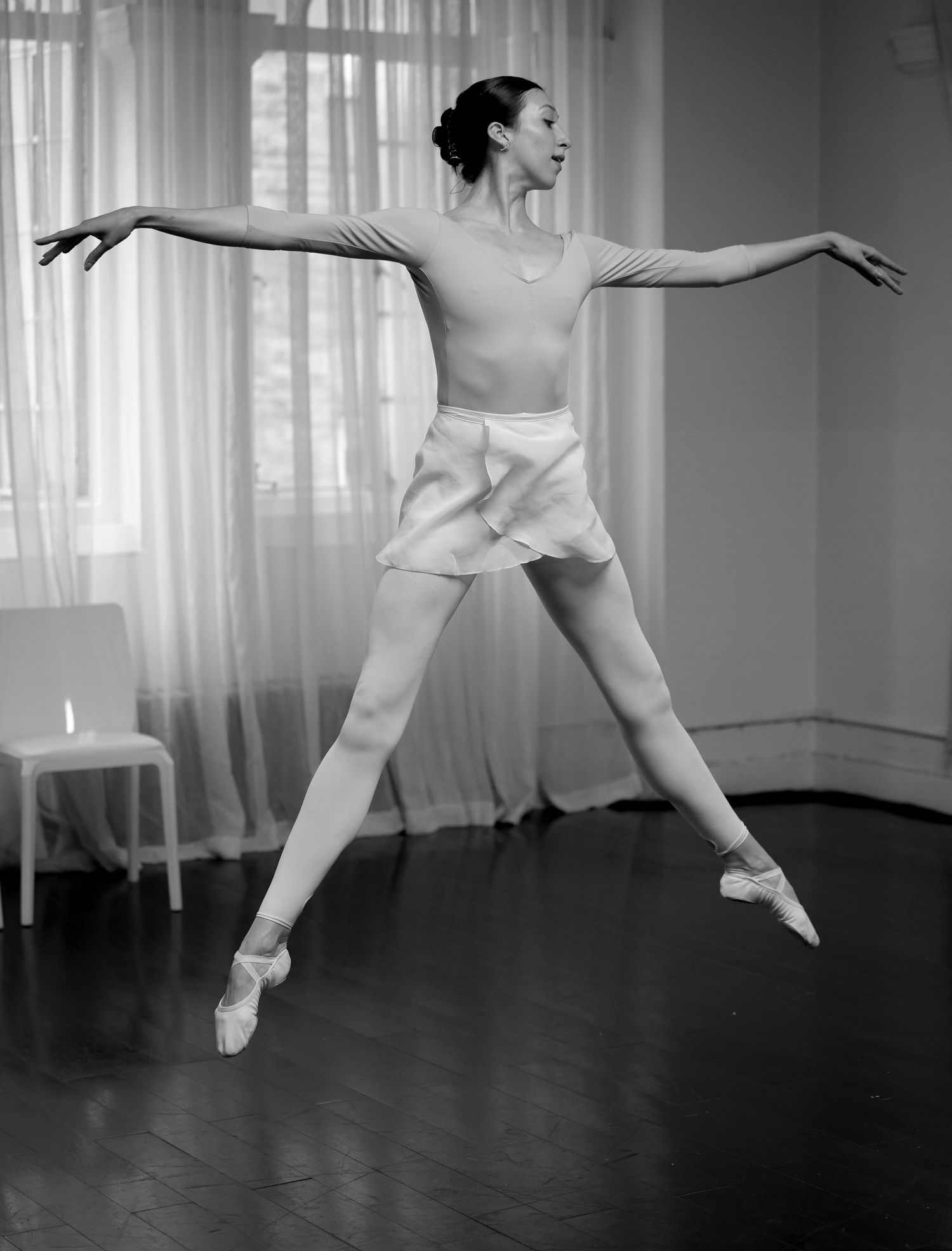 Black and white photo of a ballet dancer mid-leap in pointe shoes, arms extended gracefully in a dance studio.