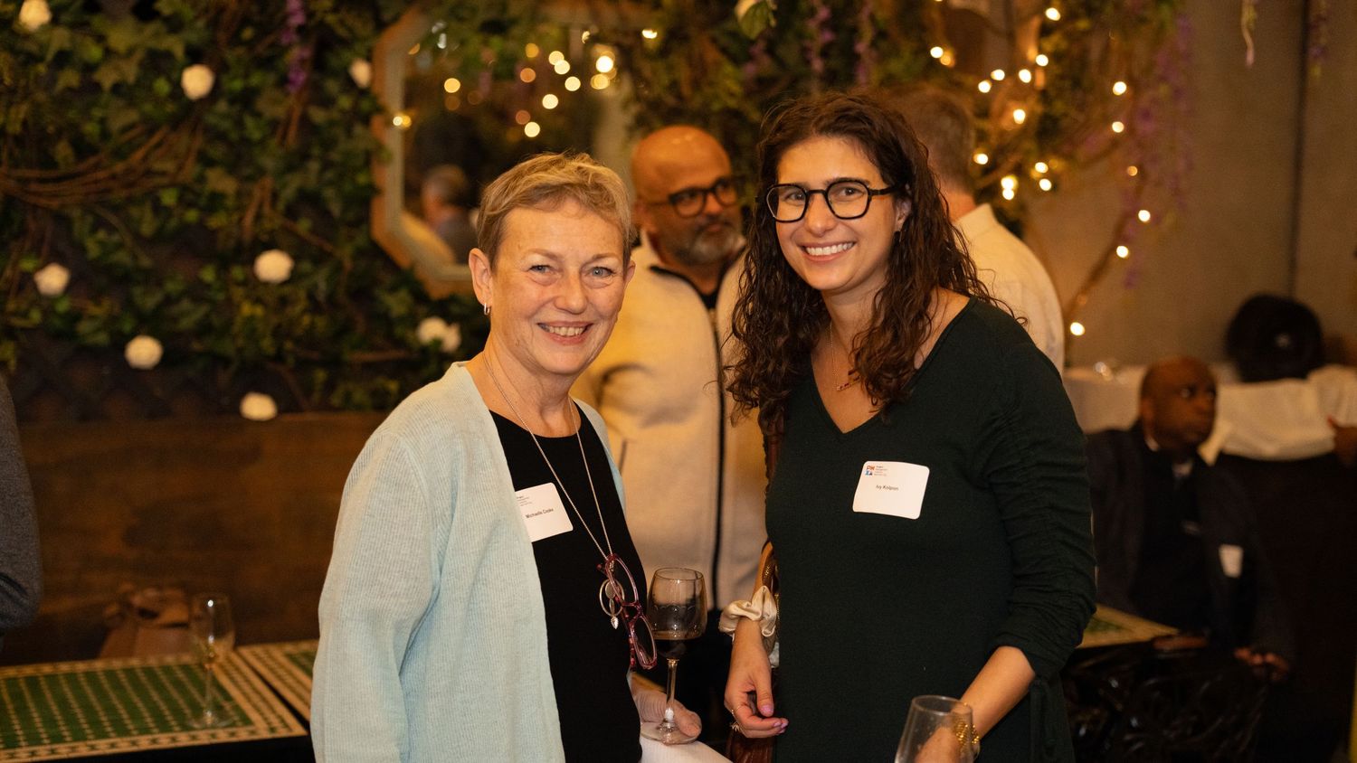 A group gathers at an evening event with warm lighting and festive decorations in the background.