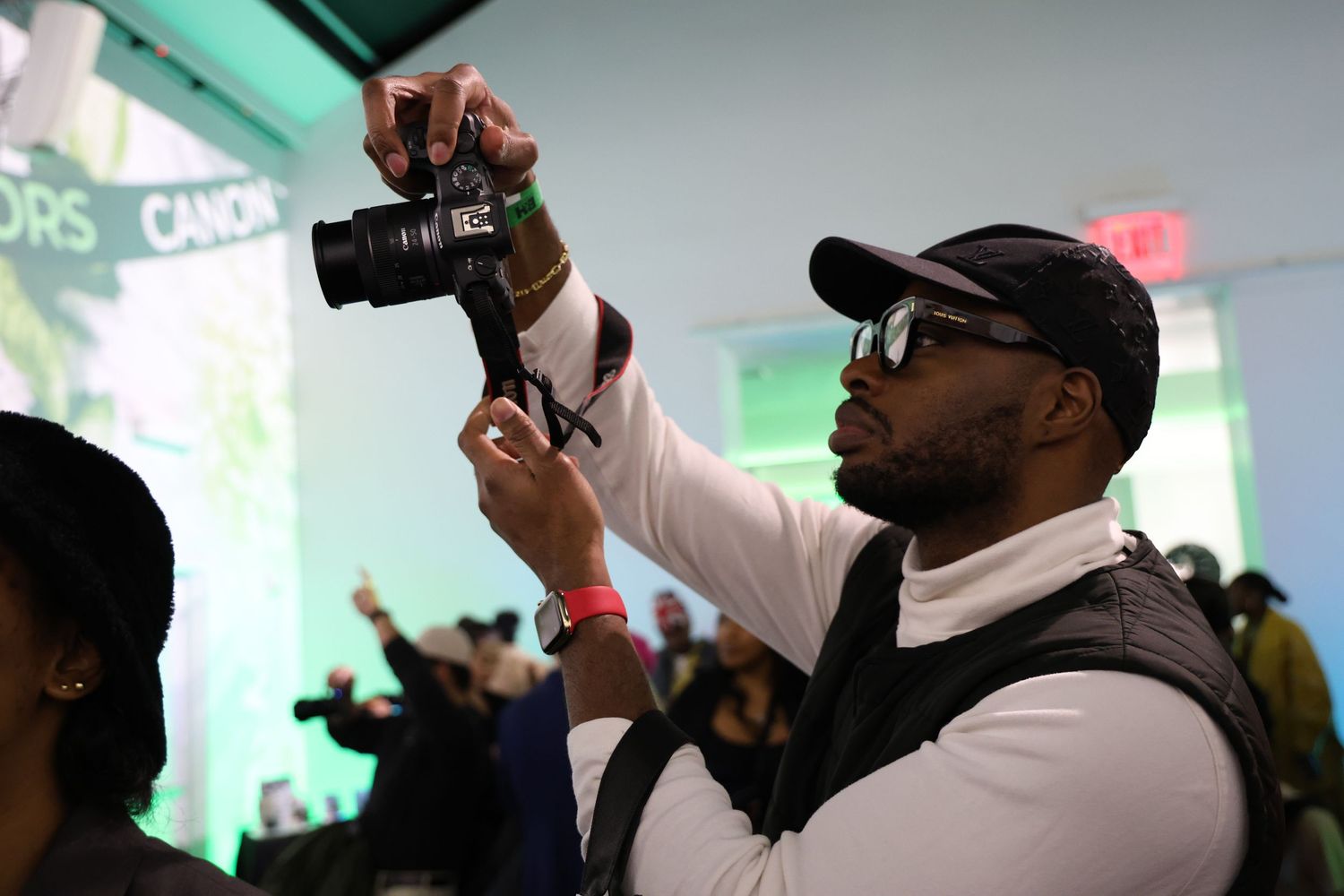 A photographer in sunglasses and a baseball cap holds up a camera during an indoor event with green lighting.