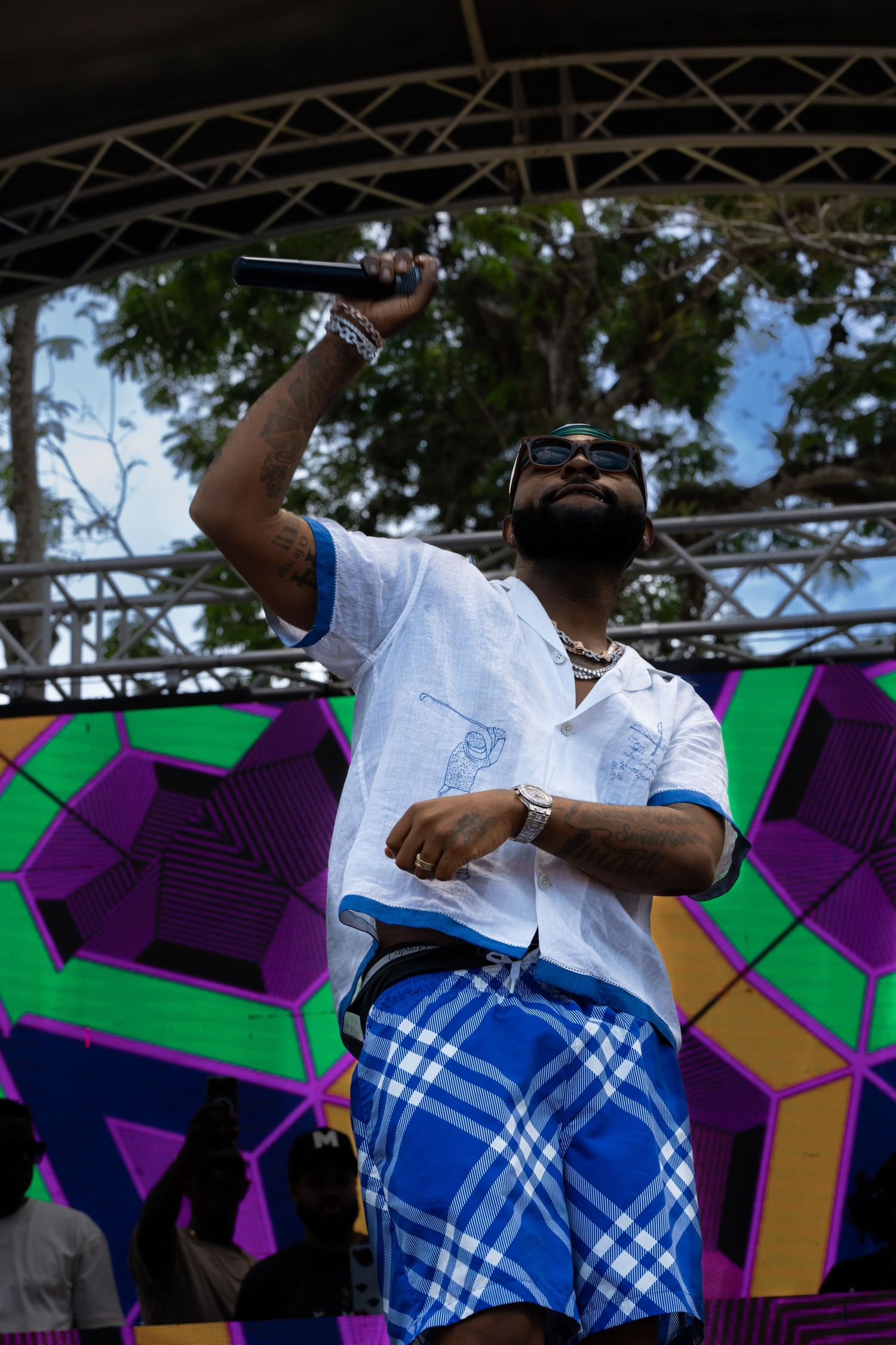 Musician on stage wearing white top and blue checkered pants against colorful geometric backdrop.