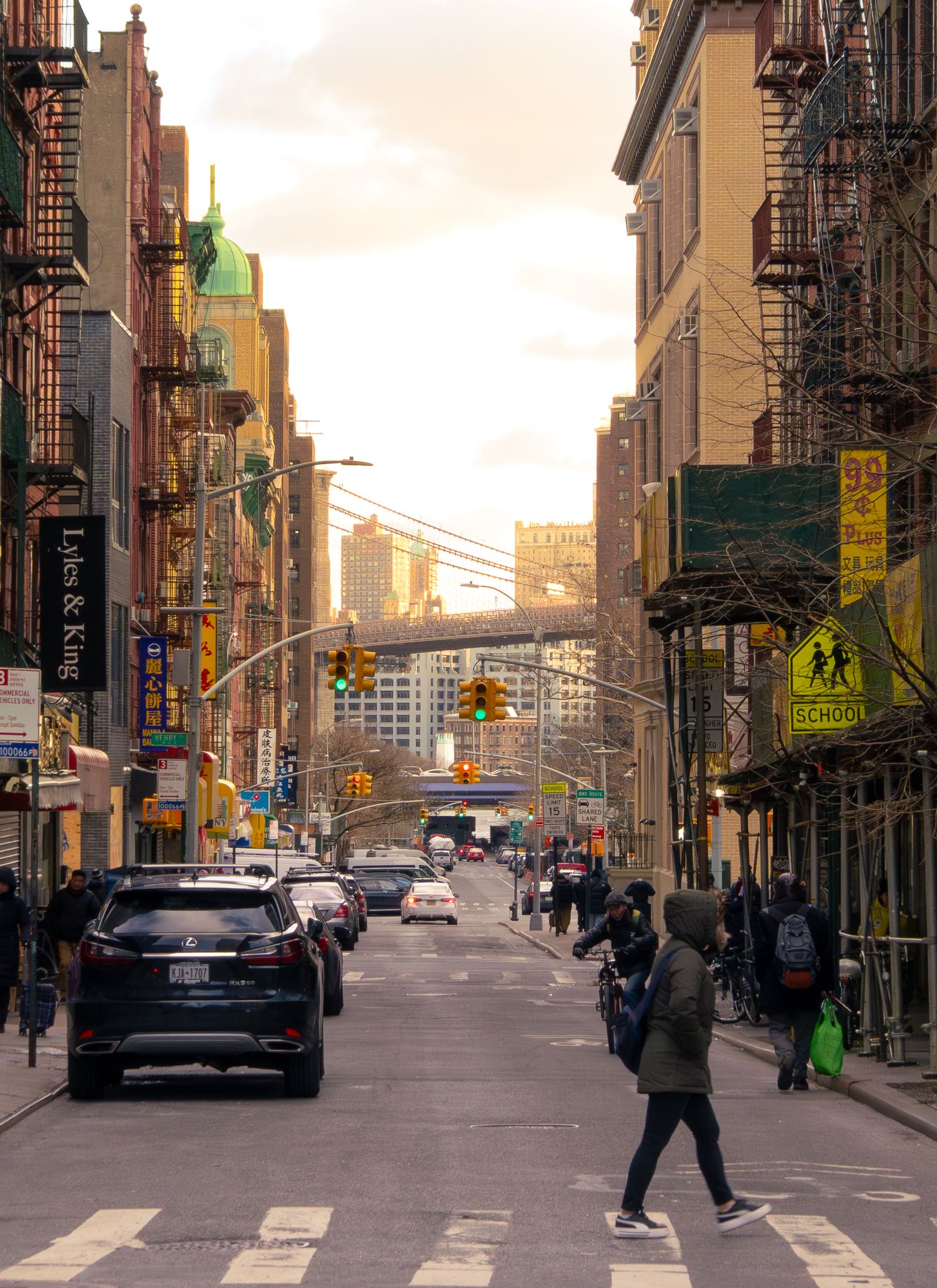 Urban street scene in New York City's Lower East Side with pedestrians crossing at sunset creating long shadows.
