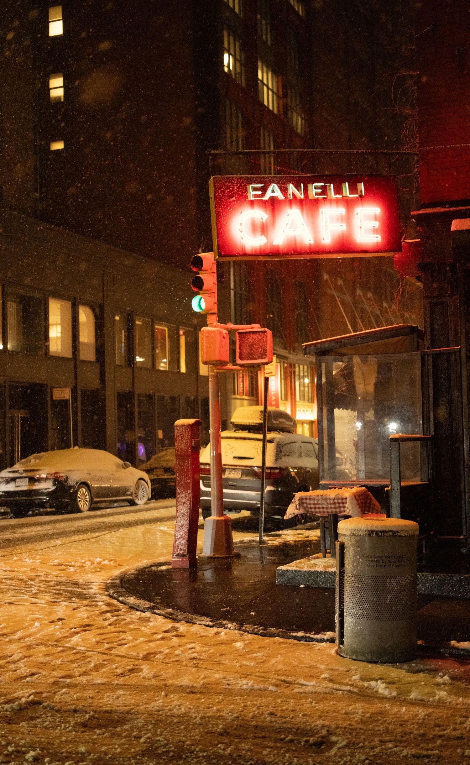 Neon red Fanelli Cafe sign glows on a snowy night in New York City with parked cars visible in the background.