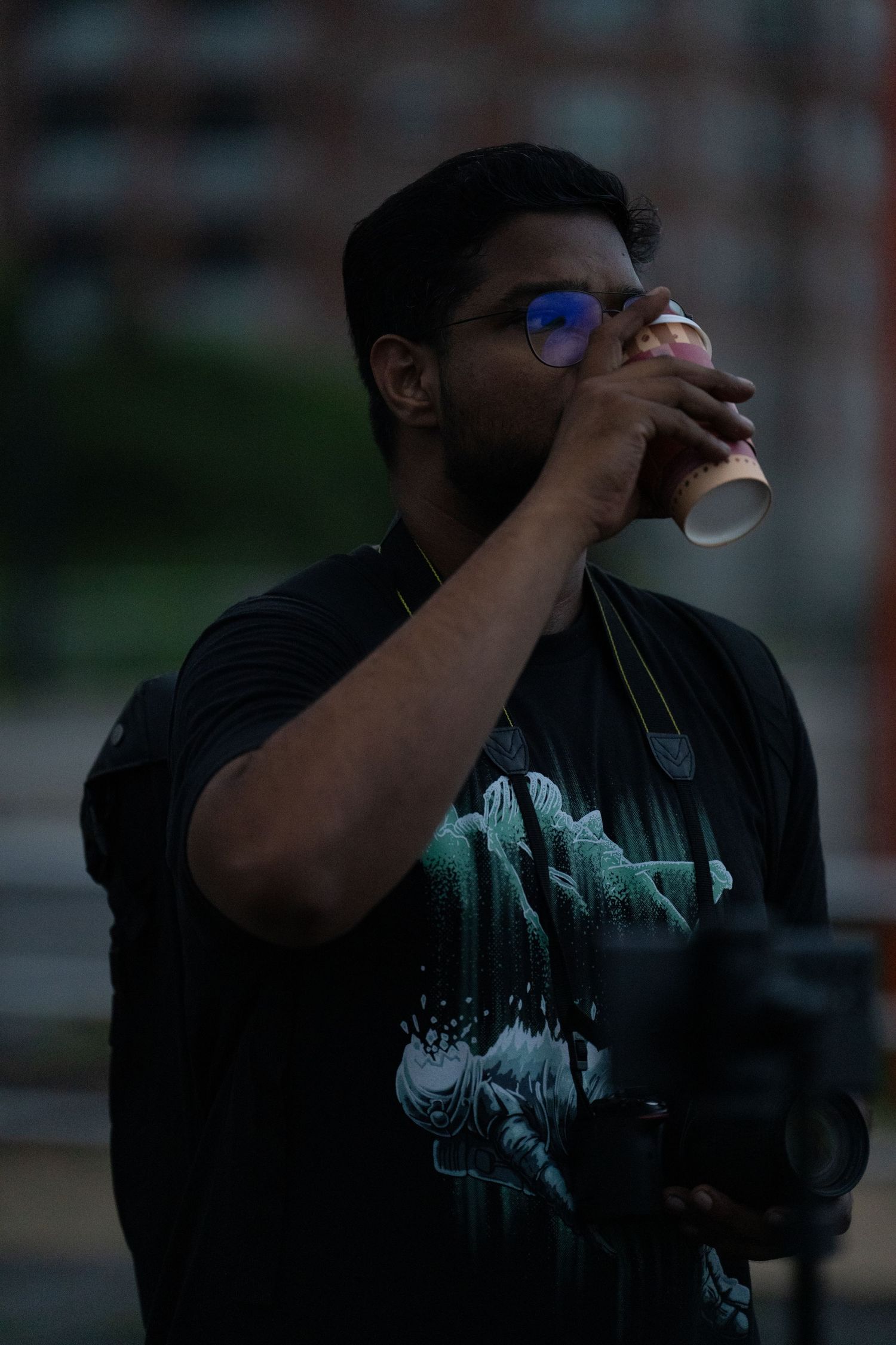 Person drinking from a coffee cup at dusk wearing a black t-shirt and blue-tinted glasses.