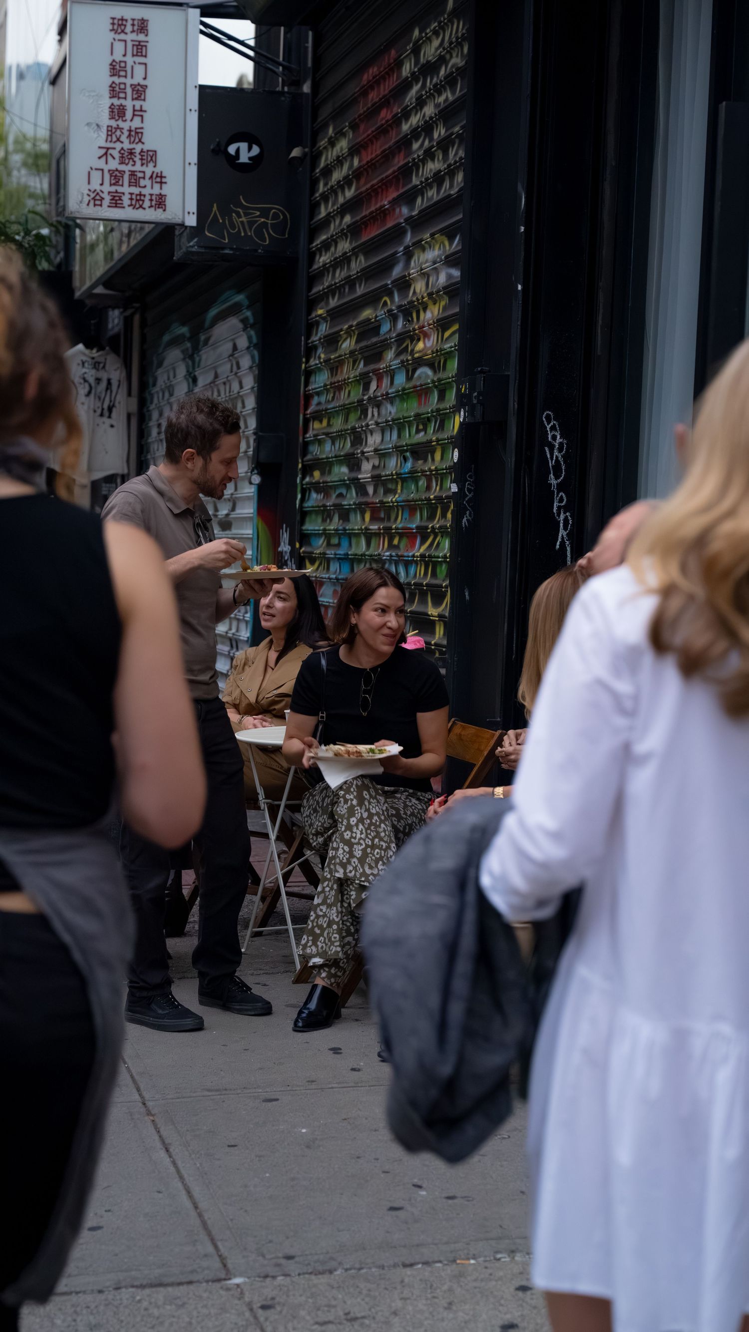 People sitting at outdoor tables along a city sidewalk with storefronts in the background.