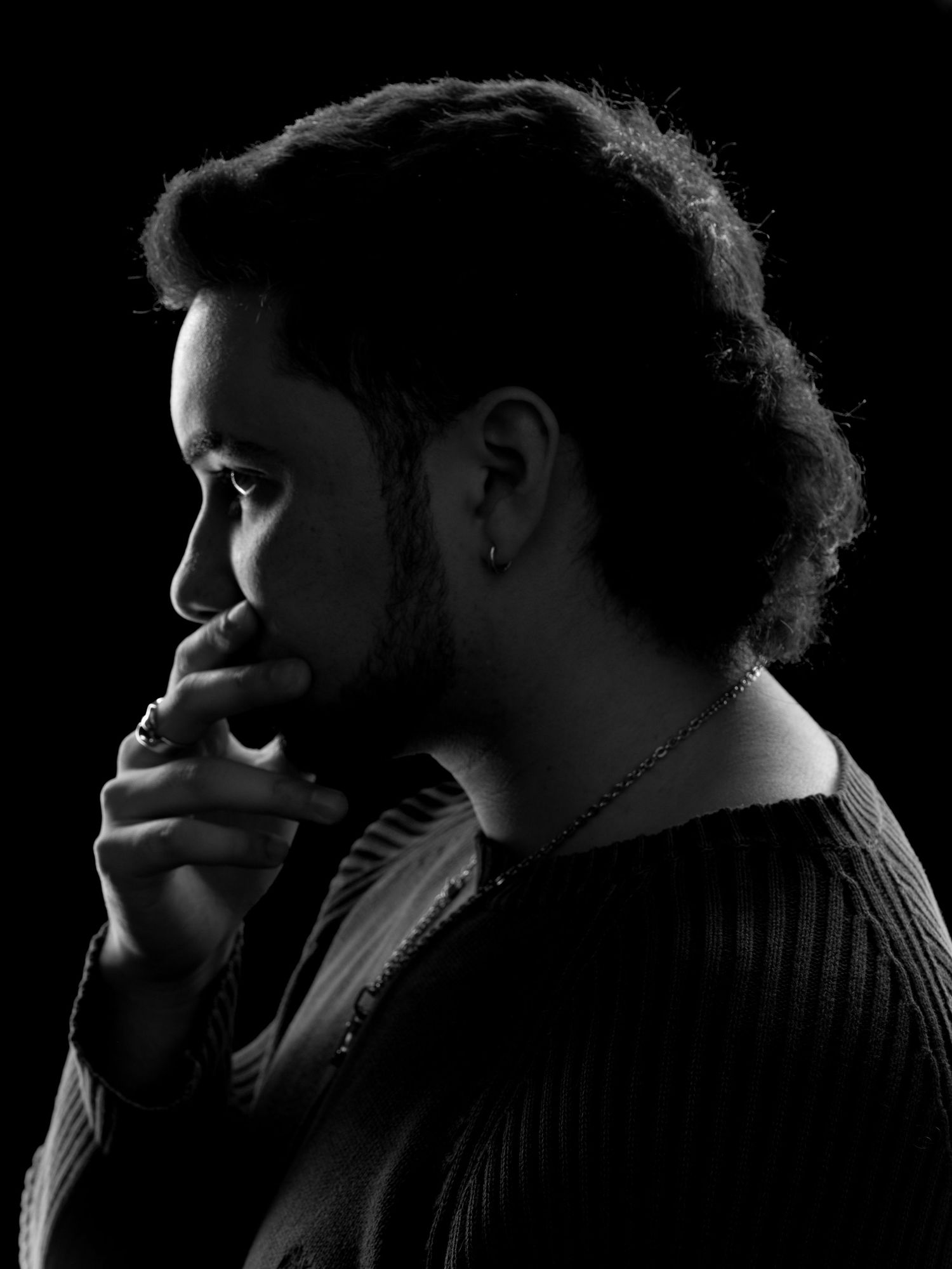 Black and white profile portrait of a thoughtful young man with curly hair, beard, and earring against dark background.
