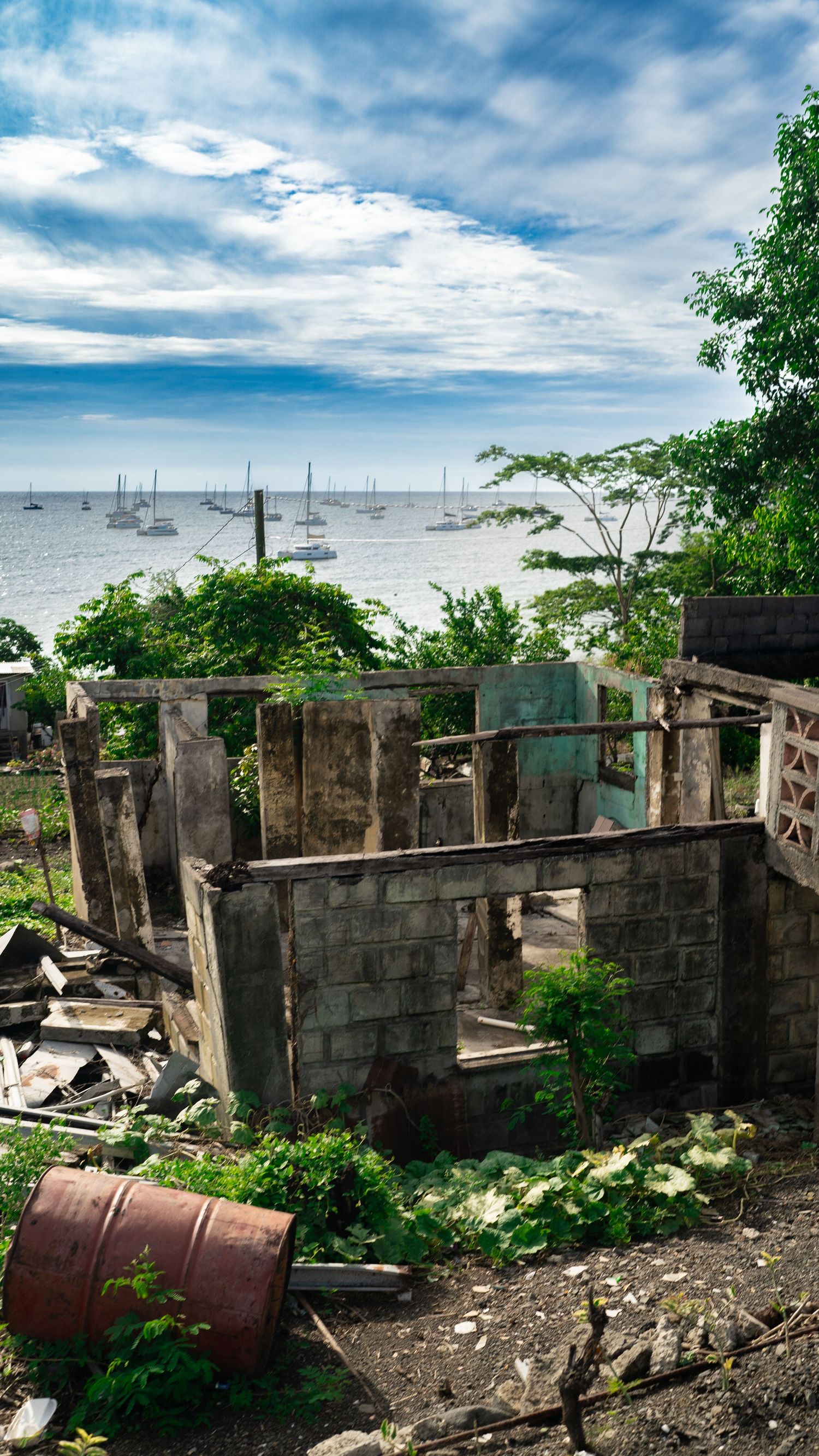 Abandoned concrete structure ruins overlooking the ocean with rusty barrels and overgrown vegetation.