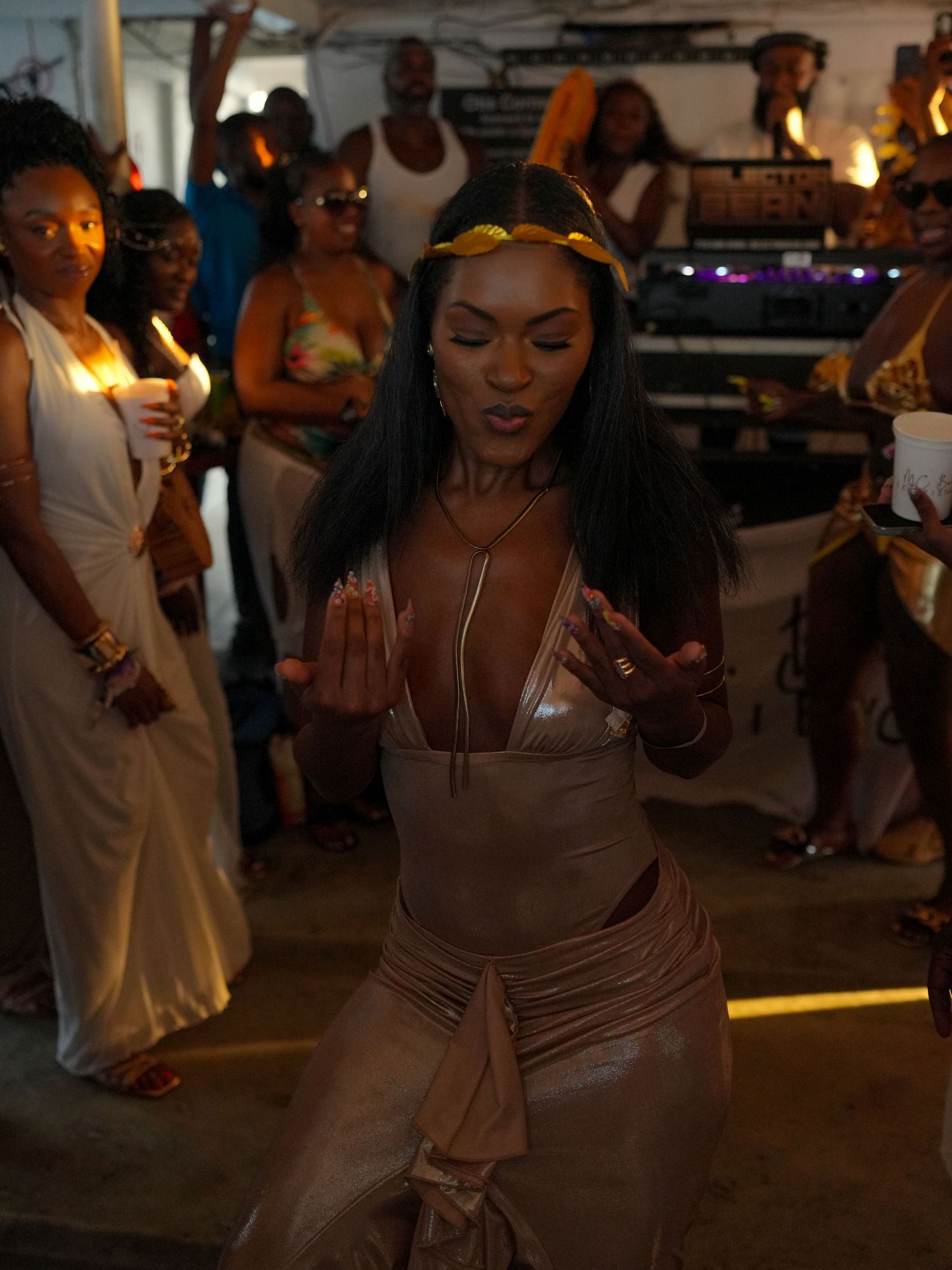 A person dances in a beige fitted dress at a dimly lit party venue with other guests visible in the background.